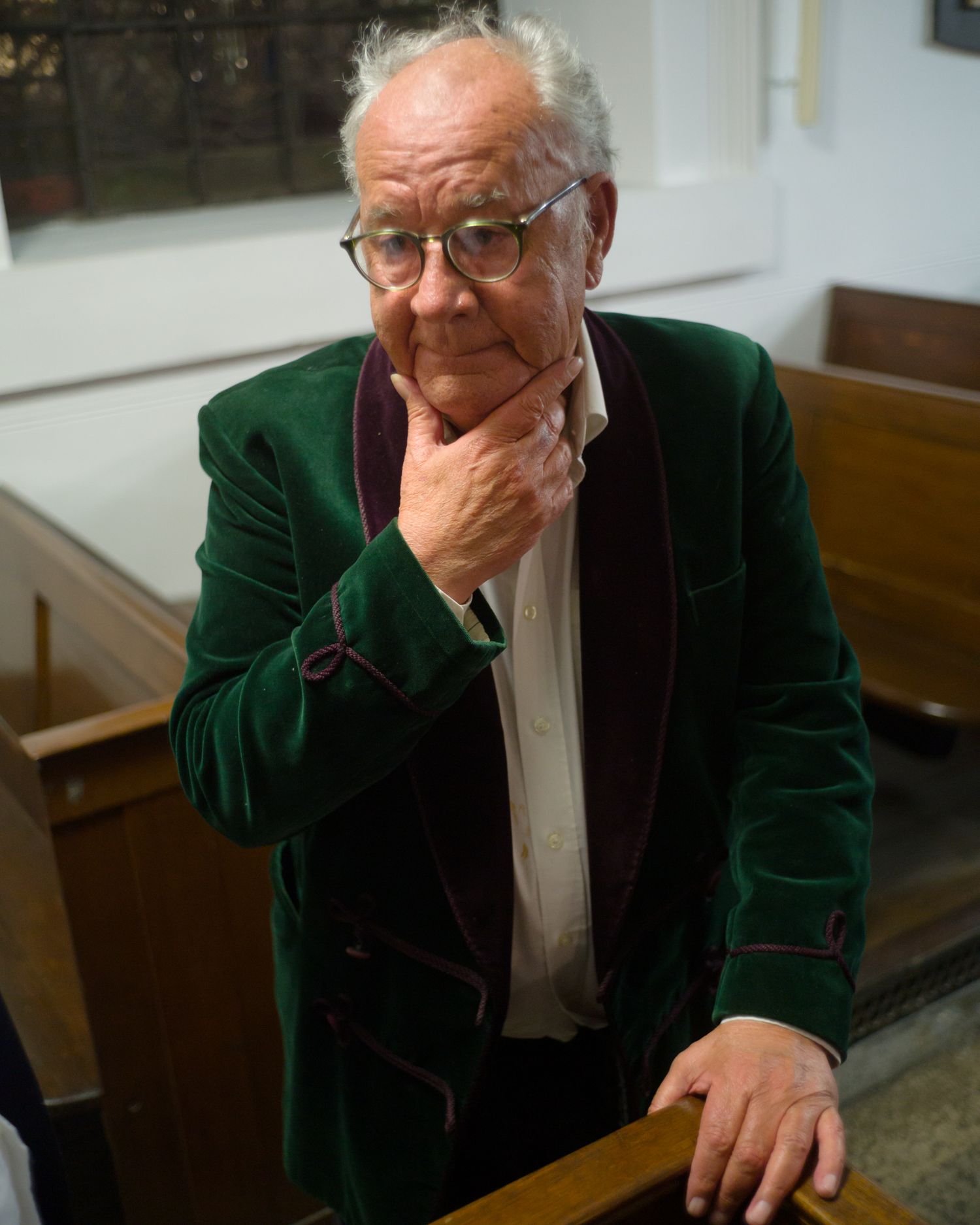 Portrait composer and guitarist Bill Loveday at his 80th birthday concert in green velvet jacket leaning on pew thoughtfully.