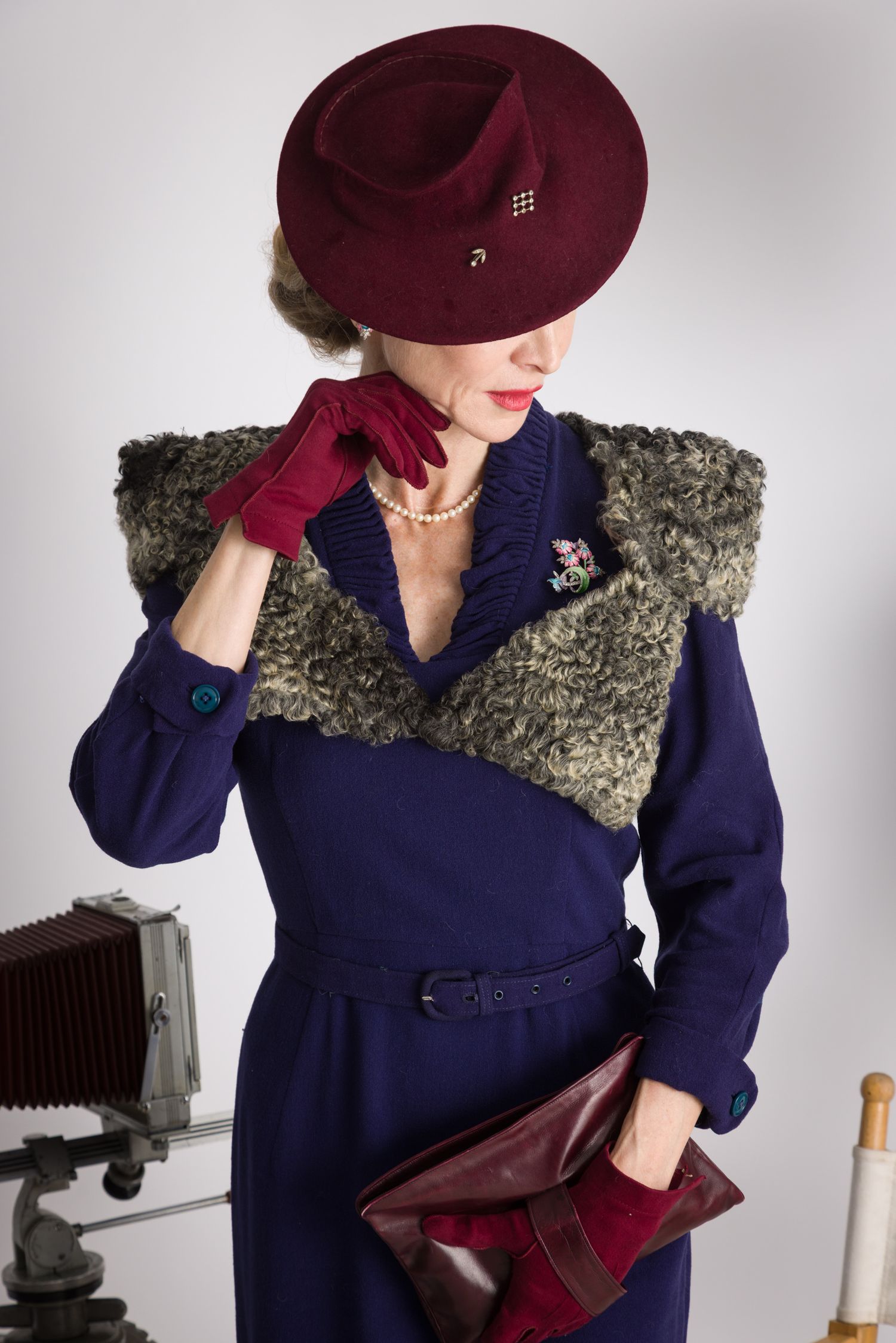 Vintage fashion photo featuring navy dress with fur collar, burgundy hat and gloves, and pearl necklace in studio setting. Goodwood Revival