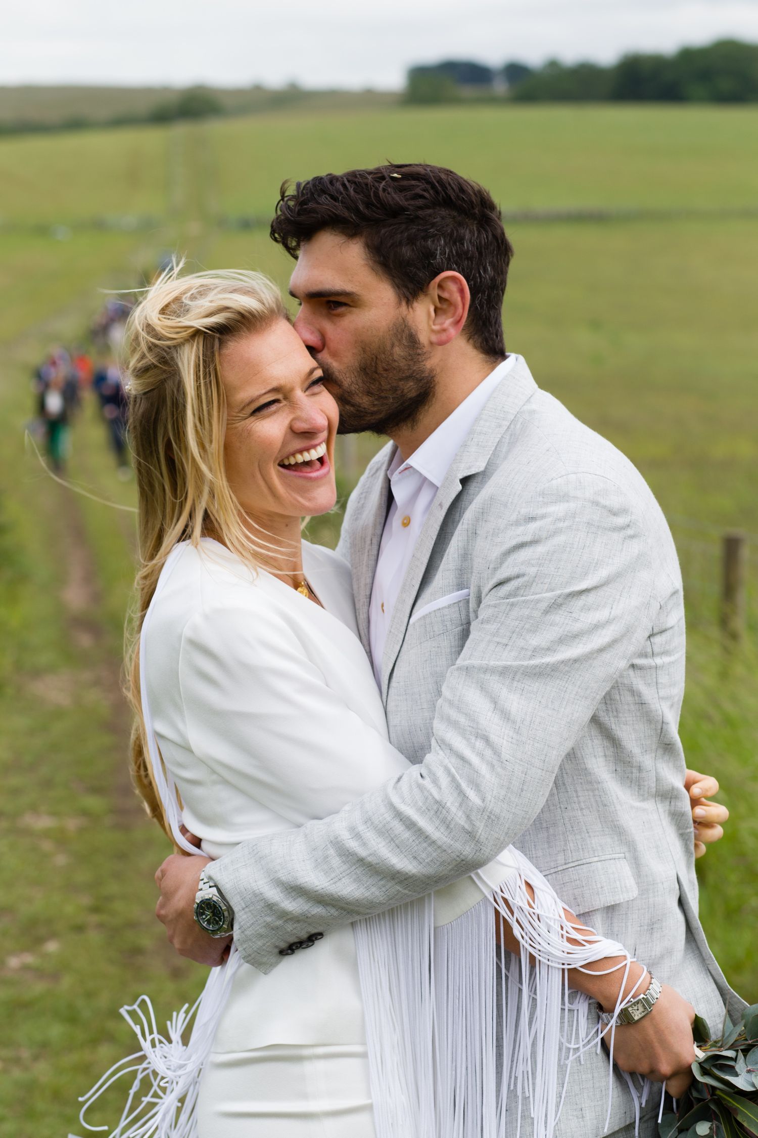 Couple in white outfits at their wedding embracing and laughing in a green field.