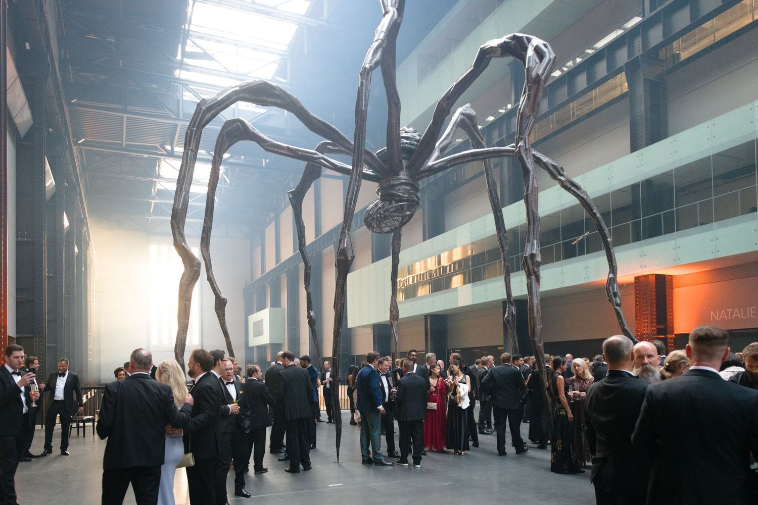 Large spider sculpture installation in a modern gallery space with formal event attendees.