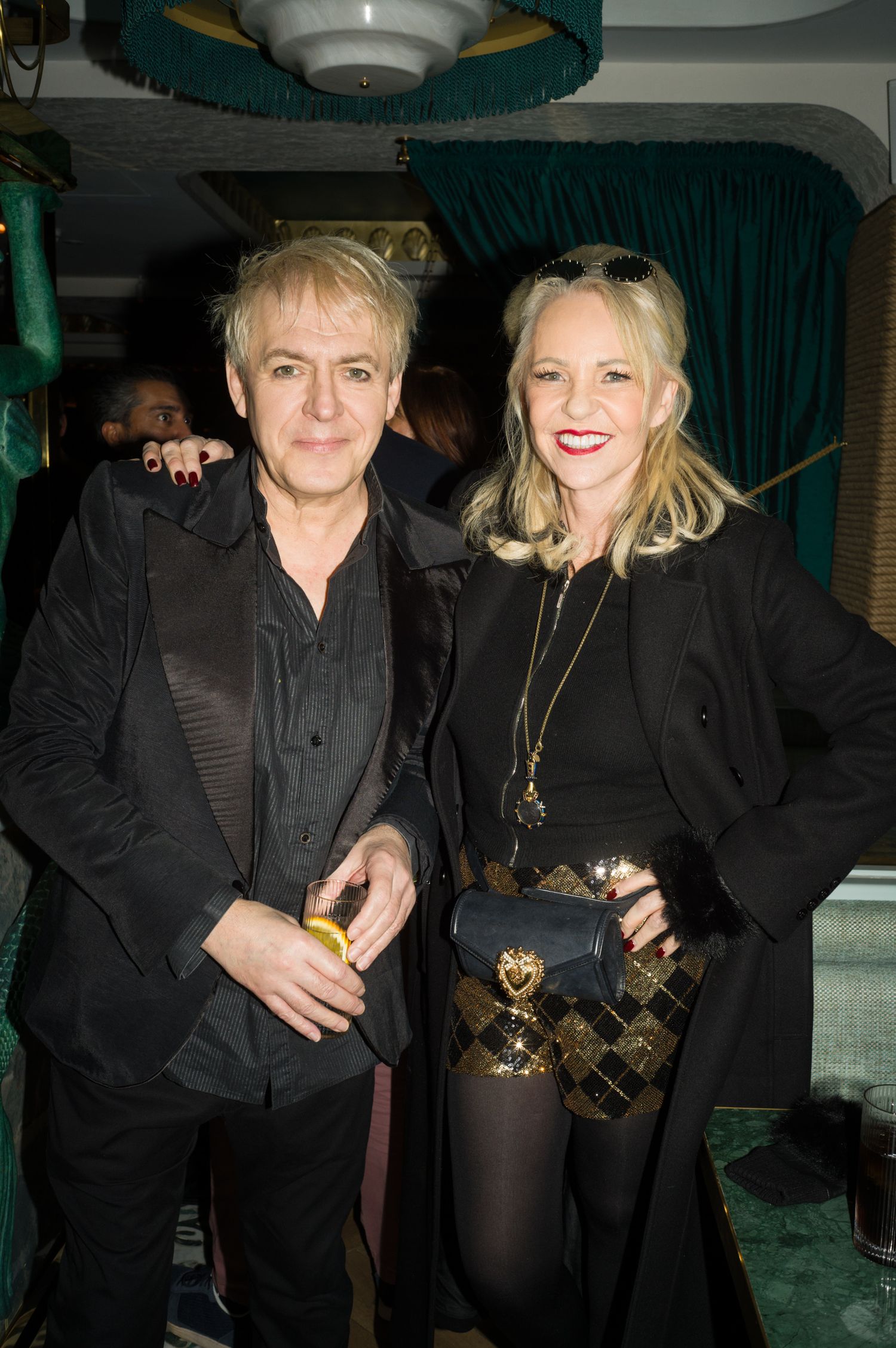Two people in black outfits posing together at an event with green curtains in the background.