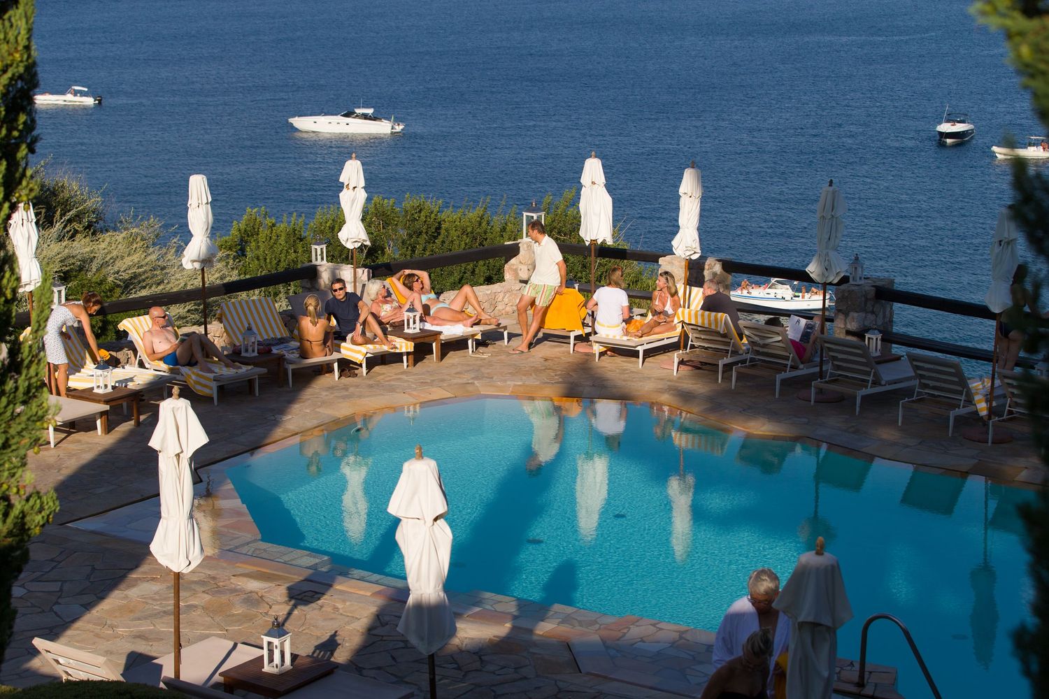 Scenic resort pool overlooking blue ocean waters with lounge chairs and umbrellas on the deck. Mediterranean summer