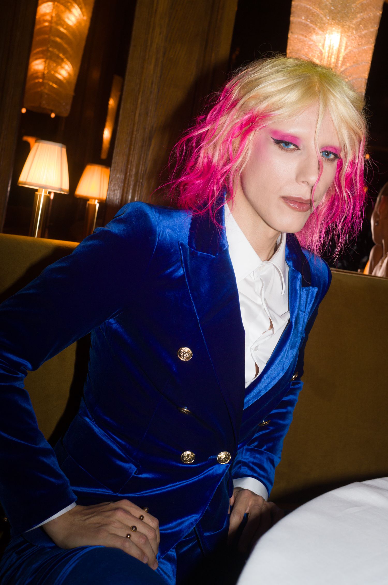 Person in royal blue velvet jacket with pink and blue hair poses against dark background.