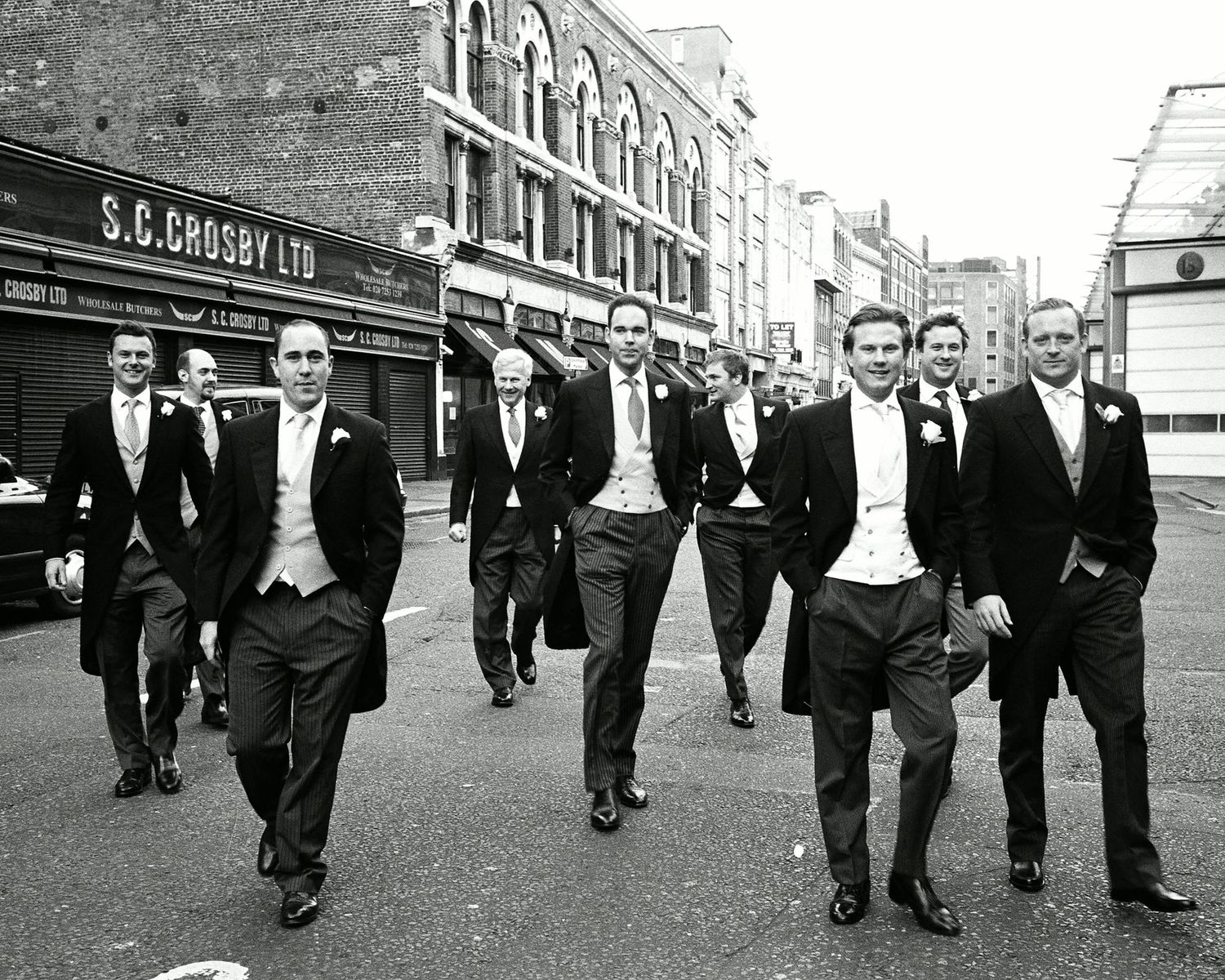 Group of people in formal wedding attire morning coats walking along a city street Smithfield Market London in b&w. Analogue Leica M