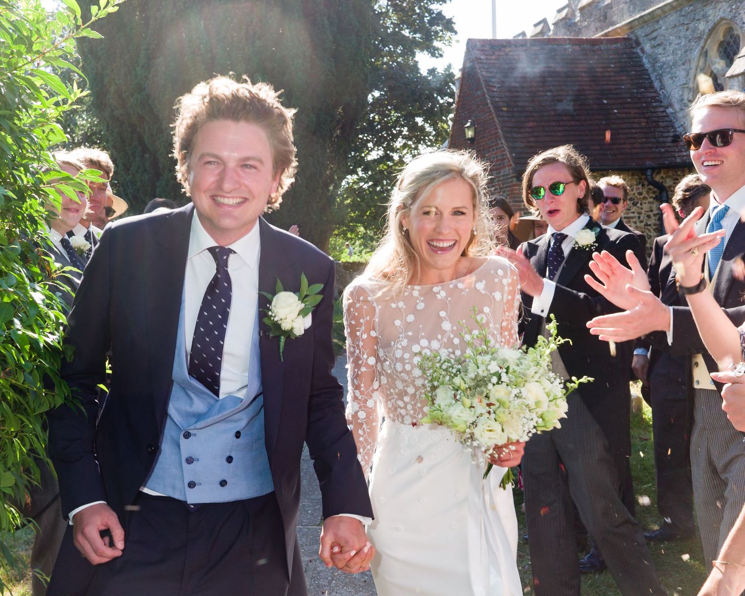 A joyful couple walks together while wedding guests celebrate in the background.