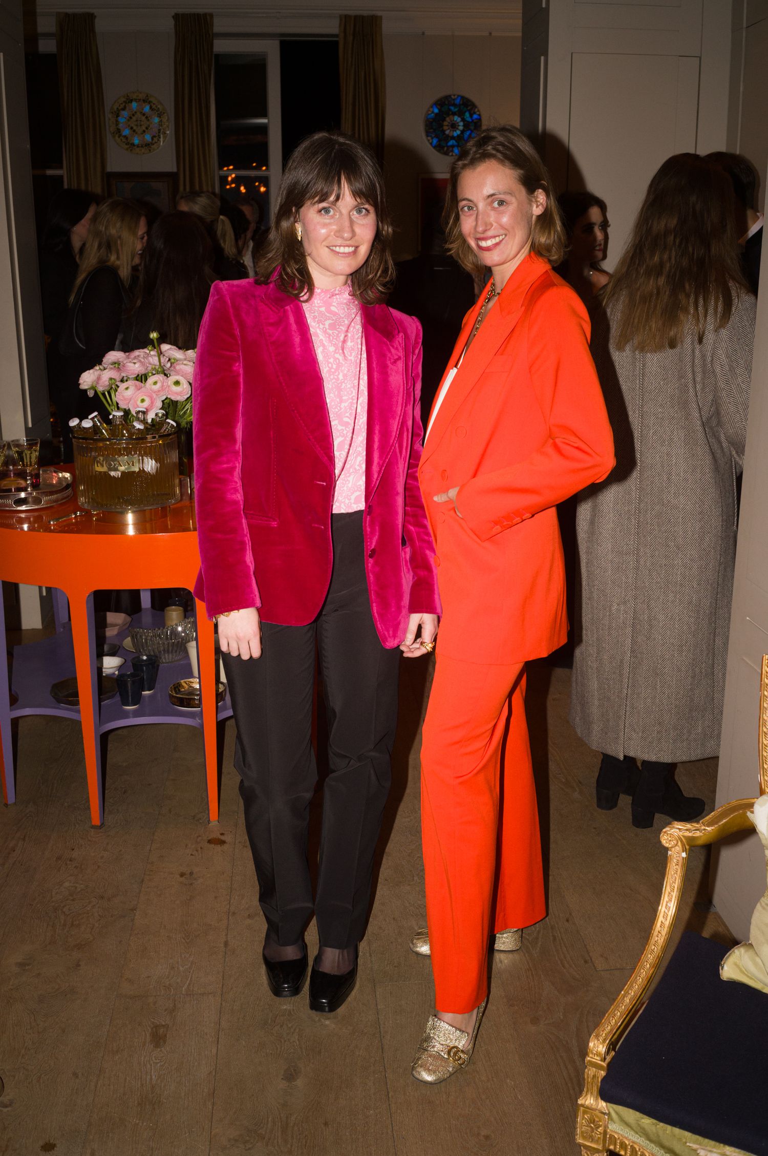 Two people posing in bright blazers, one hot pink and one orange, at an indoor gathering.