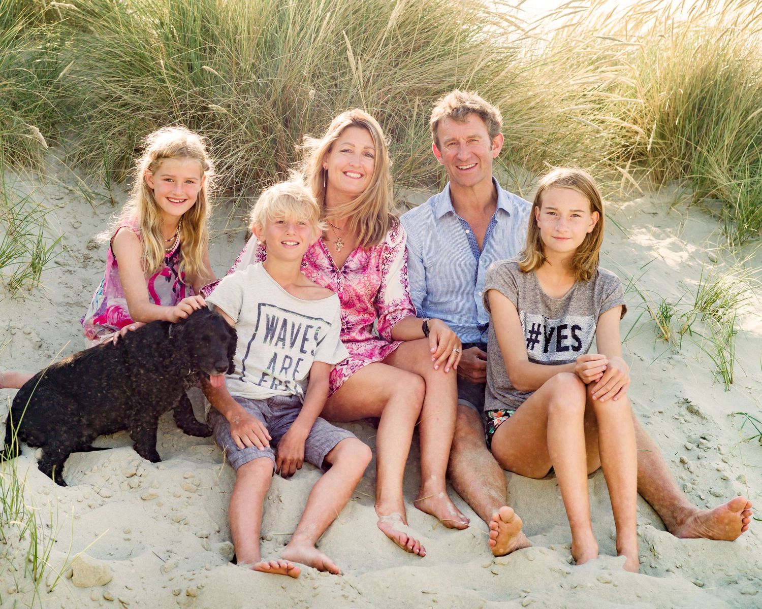 Family and black dog posing together on sandy beach dunes during sunset.