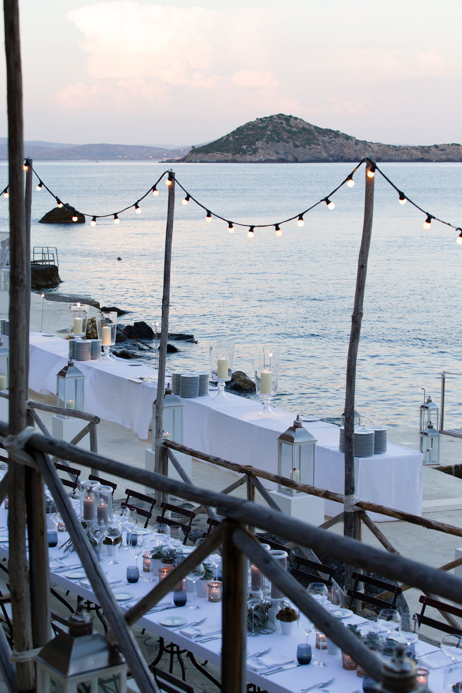 Mediterranean seaside dining setup with string lights and white tablecloths overlooking an island at dusk.
