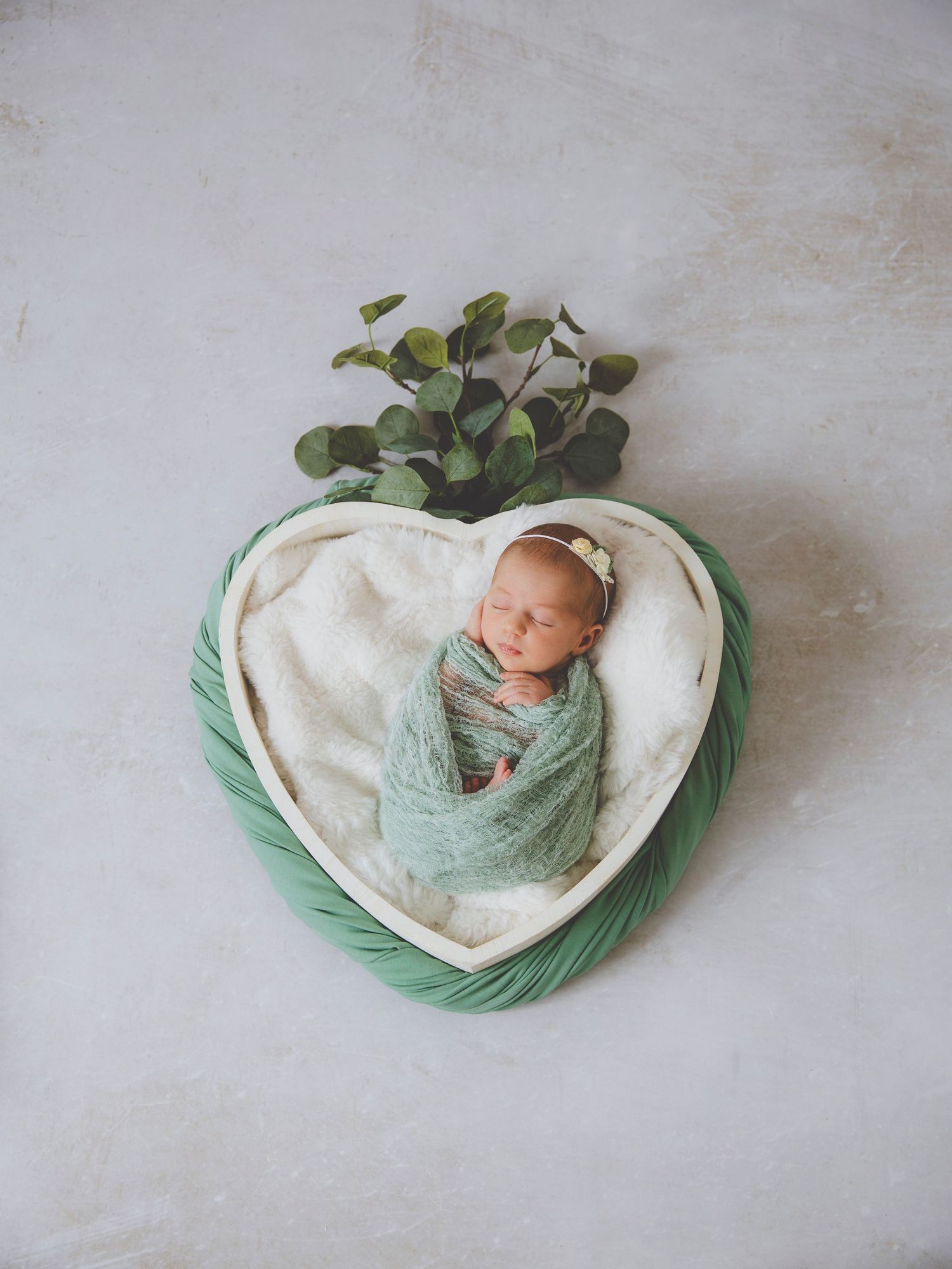 Recién nacida dormida, envuelta en una tela verde, dentro de un cuenco con forma de corazón decorado con hojas de eucalipto.