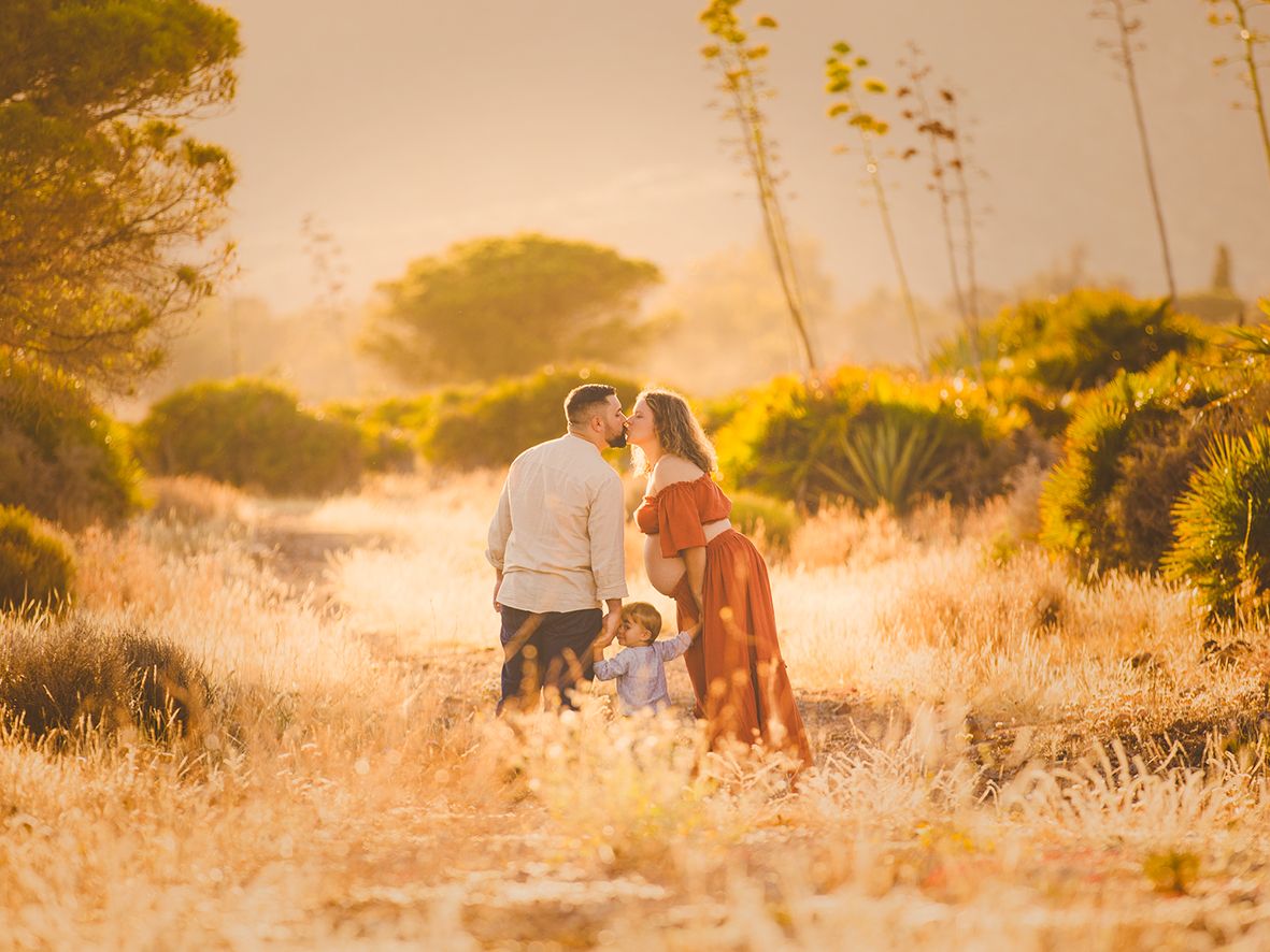 Una pareja embarazada se da un beso al atardecer con su hijo pequeño en los campos de Genoveses bañados por el sol del atardecer.