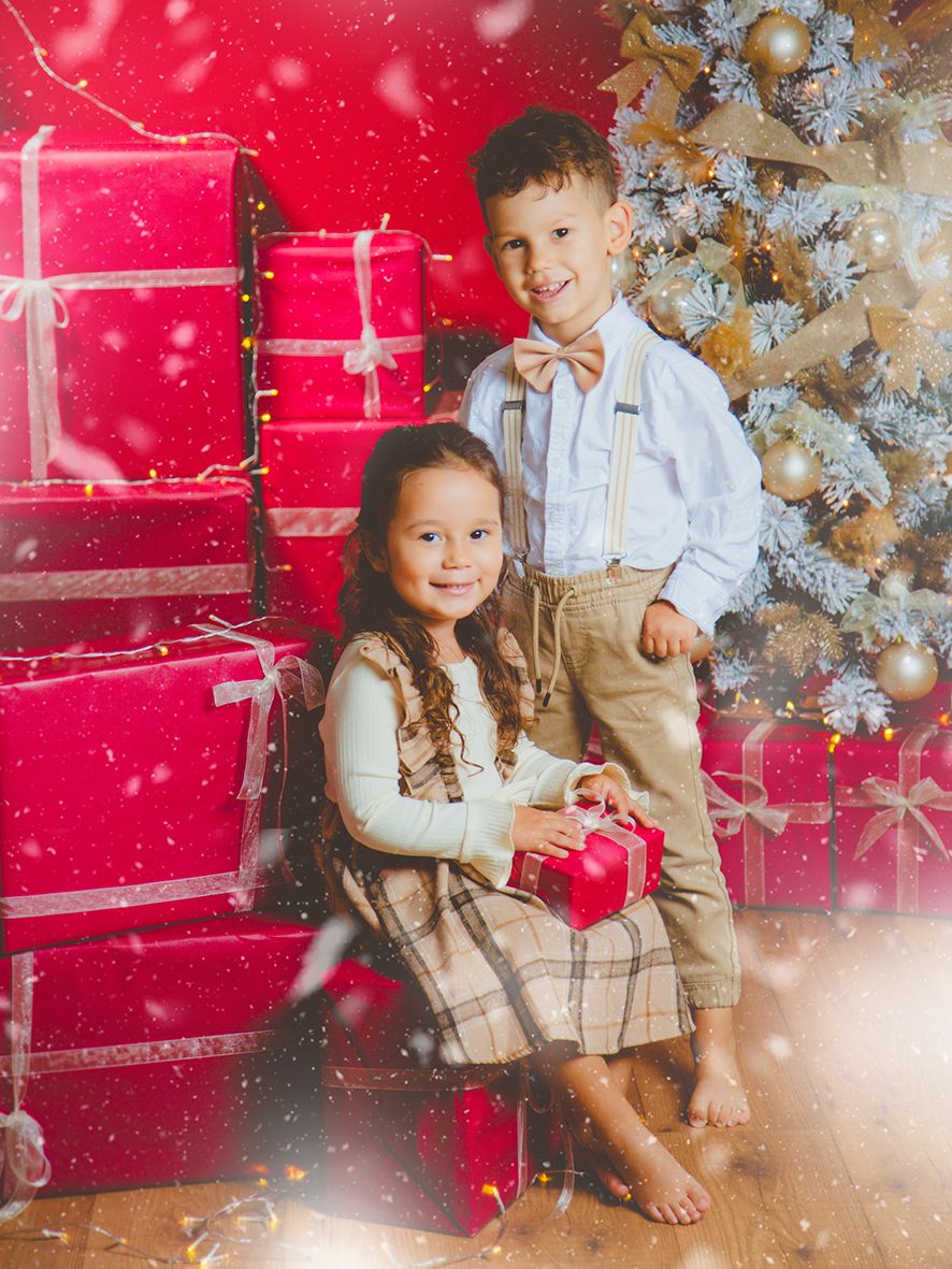 Unos niños posan sentados juntos frente a una pared roja y un árbol de Navidad blanco decorado con adornos dorados.