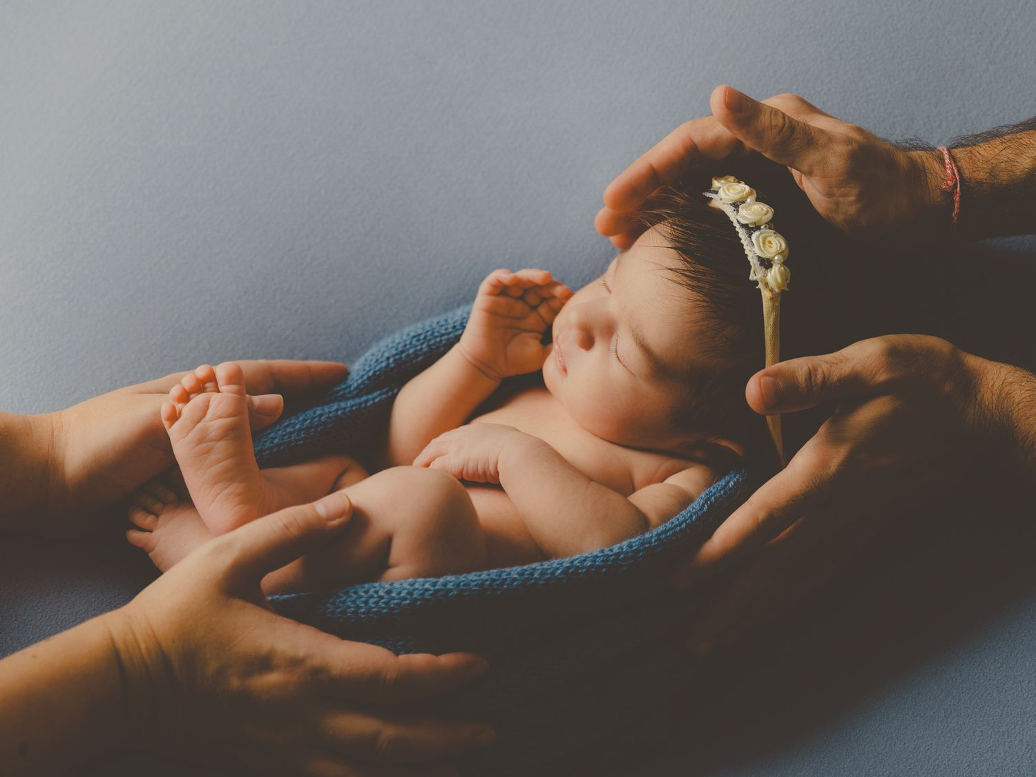 Bebé recién nacido con corona de flores durmiendo envuelto en una manta azul, acunado por las manos de sus padres.
