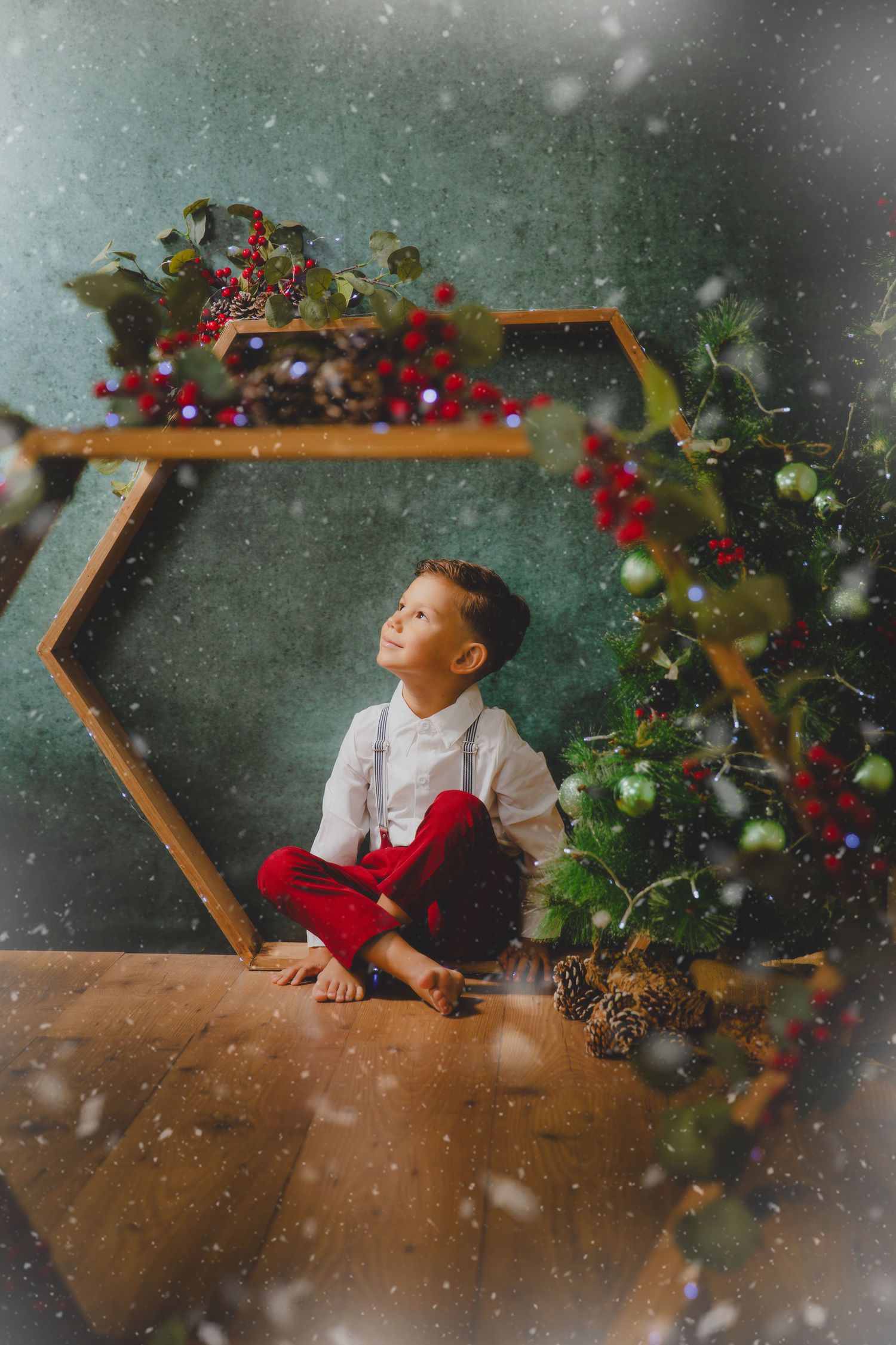 Un niño con pantalón rojo está sentado dentro de un marco hexagonal de madera decorado con ramas navideñas y bayas.