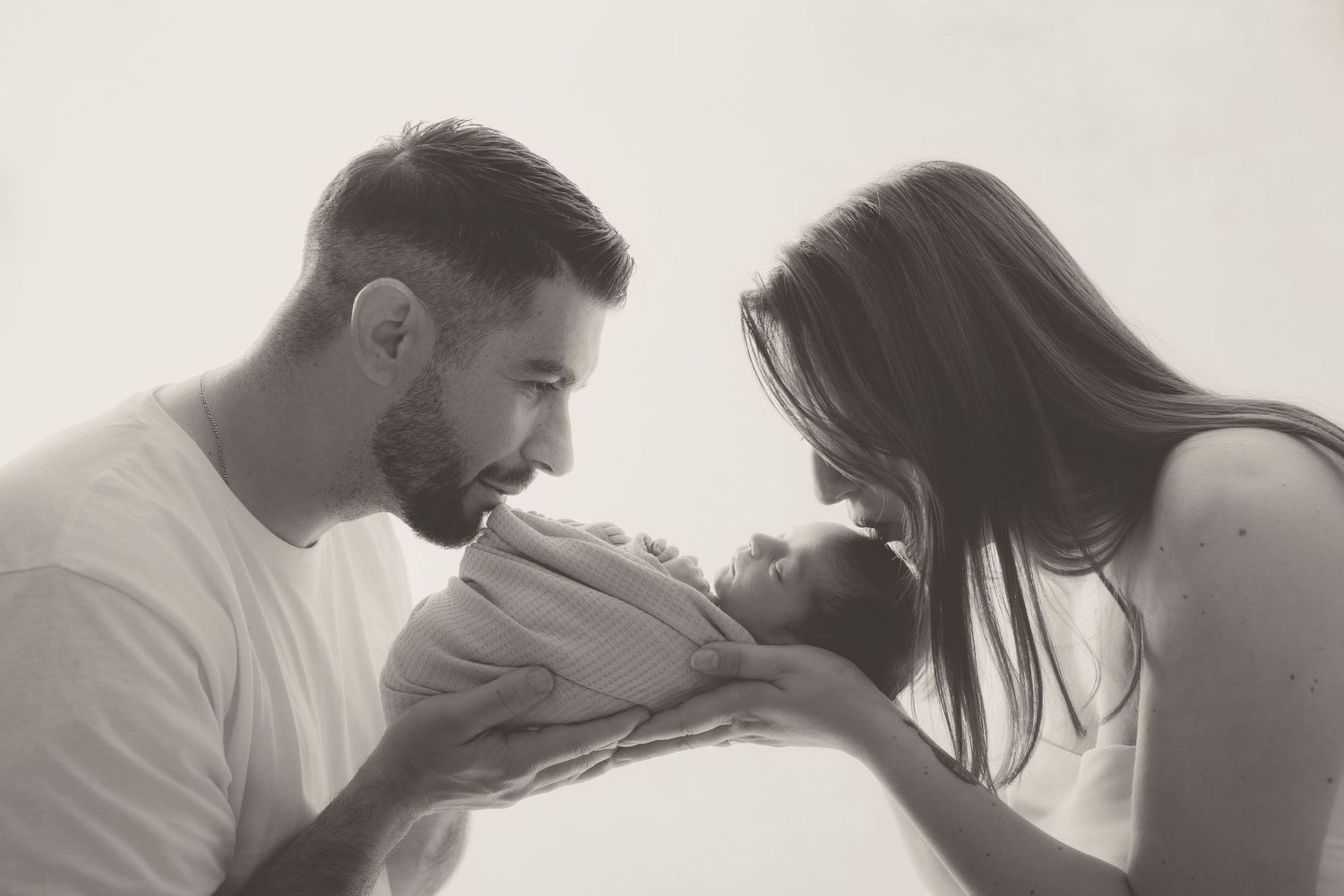 Fotografía en blanco y negro de unos padres besando tiernamente a su bebé recién nacido envuelto en una tela.