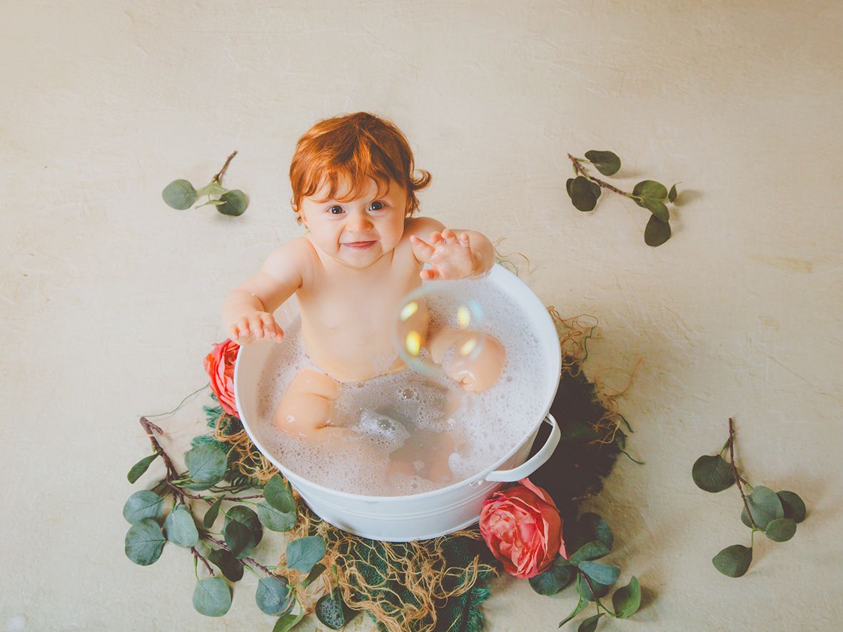 Adorable bebé pelirroja en su sesión de primer cumpleaños en una bañera de agua con hojas verdes y flores de decorado.