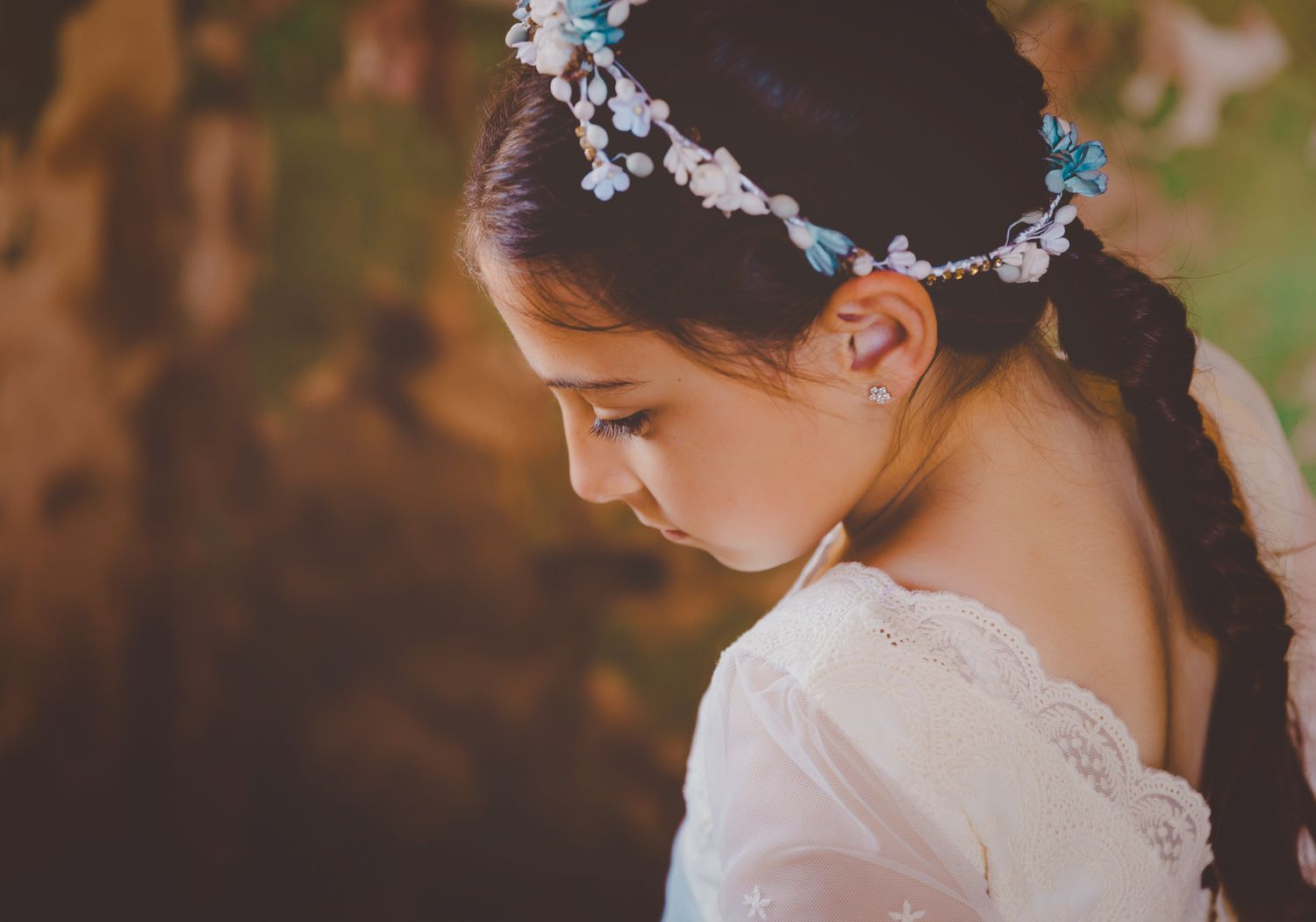 Perfil de una niña con vestido blanco de comunión, diadema floral azul y cabello trenzado en un entorno natural.