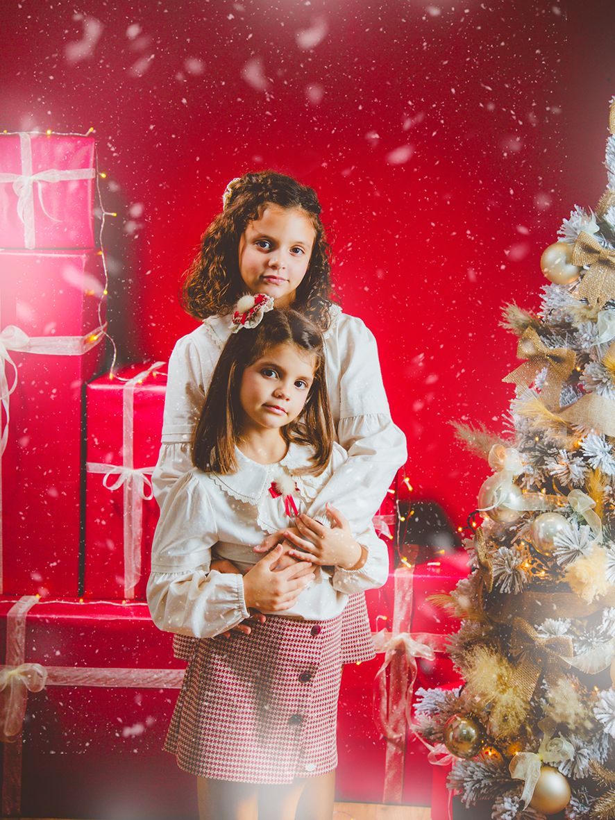 Dos hermanas posando para su sesión de fotos al lado de un árbol de Navidad y cajas de regalos en un fondo rojo.