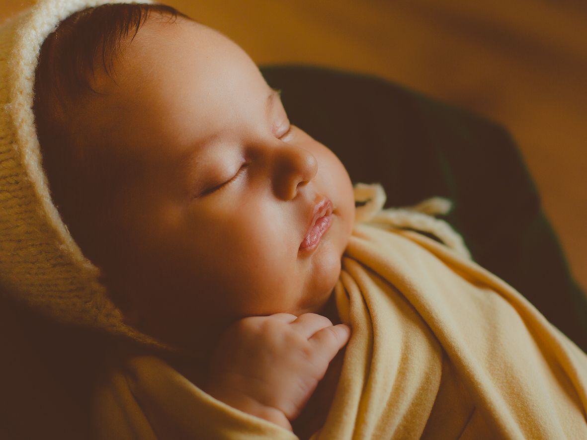 Un bebé recién nacido, tranquilo y envuelto en una suave tela color crema con gorrito de lana, duerme bajo la cálida luz del estudio.