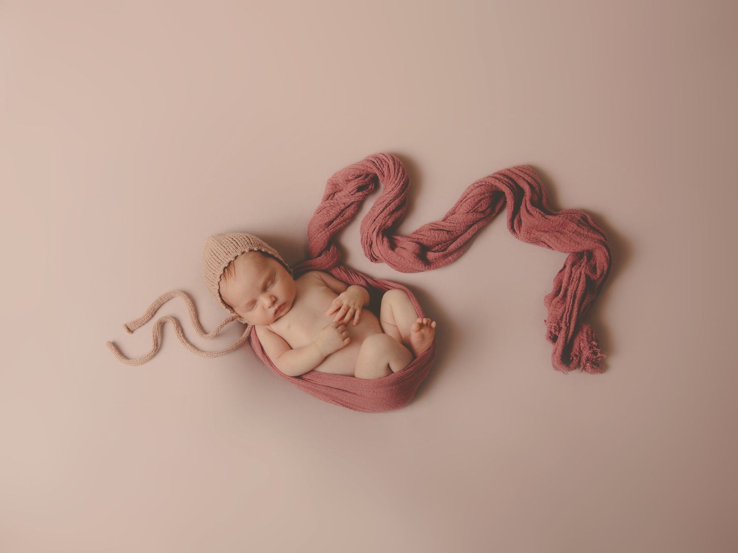 Bebé recién nacido dormido en su sesión de fotos, envuelto en una tela color rosa empolvado con un gorrito a juego sobre un fondo rosa.