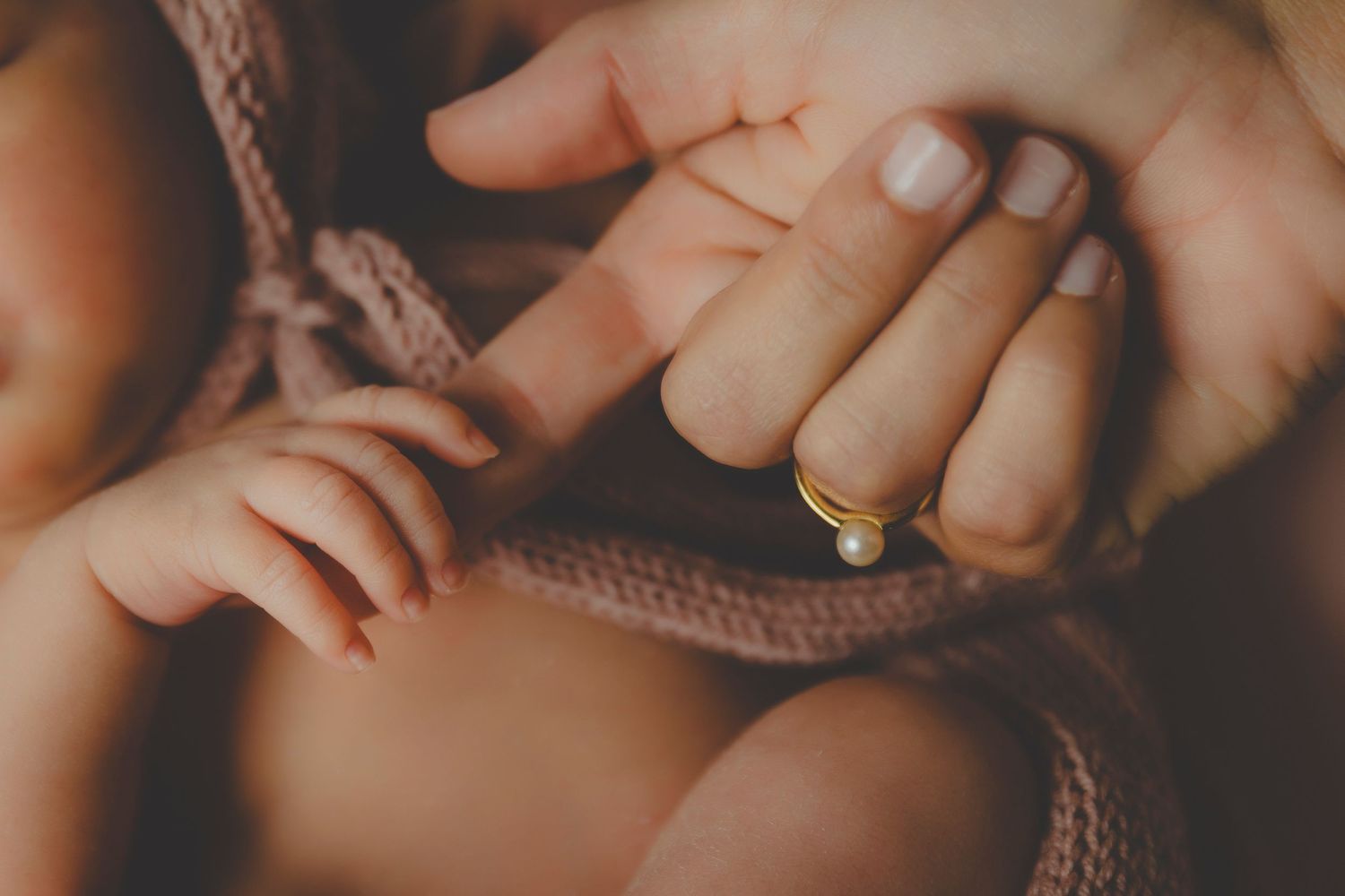 Primer plano de los dedos de un bebé recién nacido sujetando suavemente la mano de su madre, que lleva un anillo dorado con una perla.