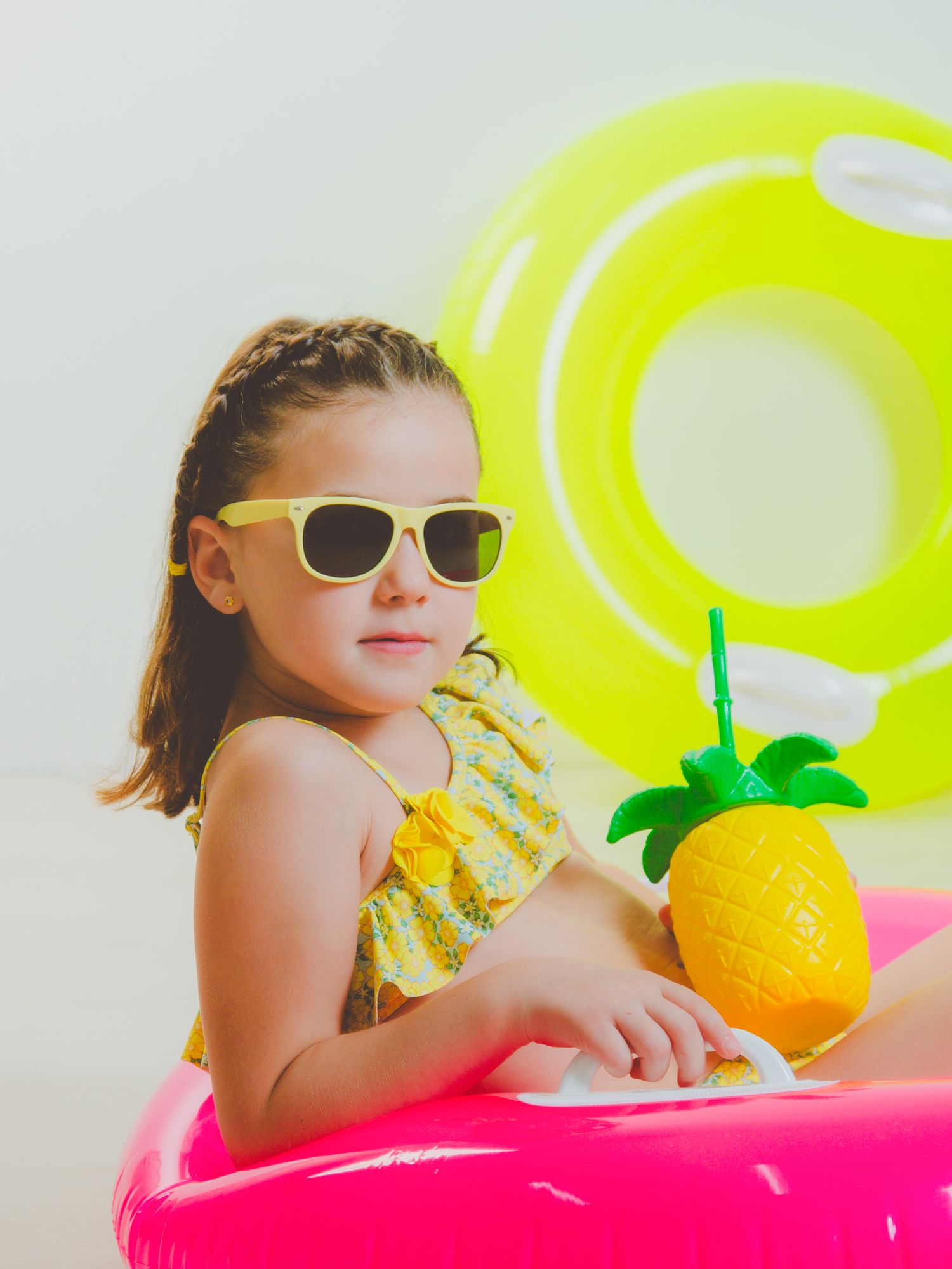 Niña en su sesión de verano, con gafas de sol en un flotador fucsia y con vaso de piña.