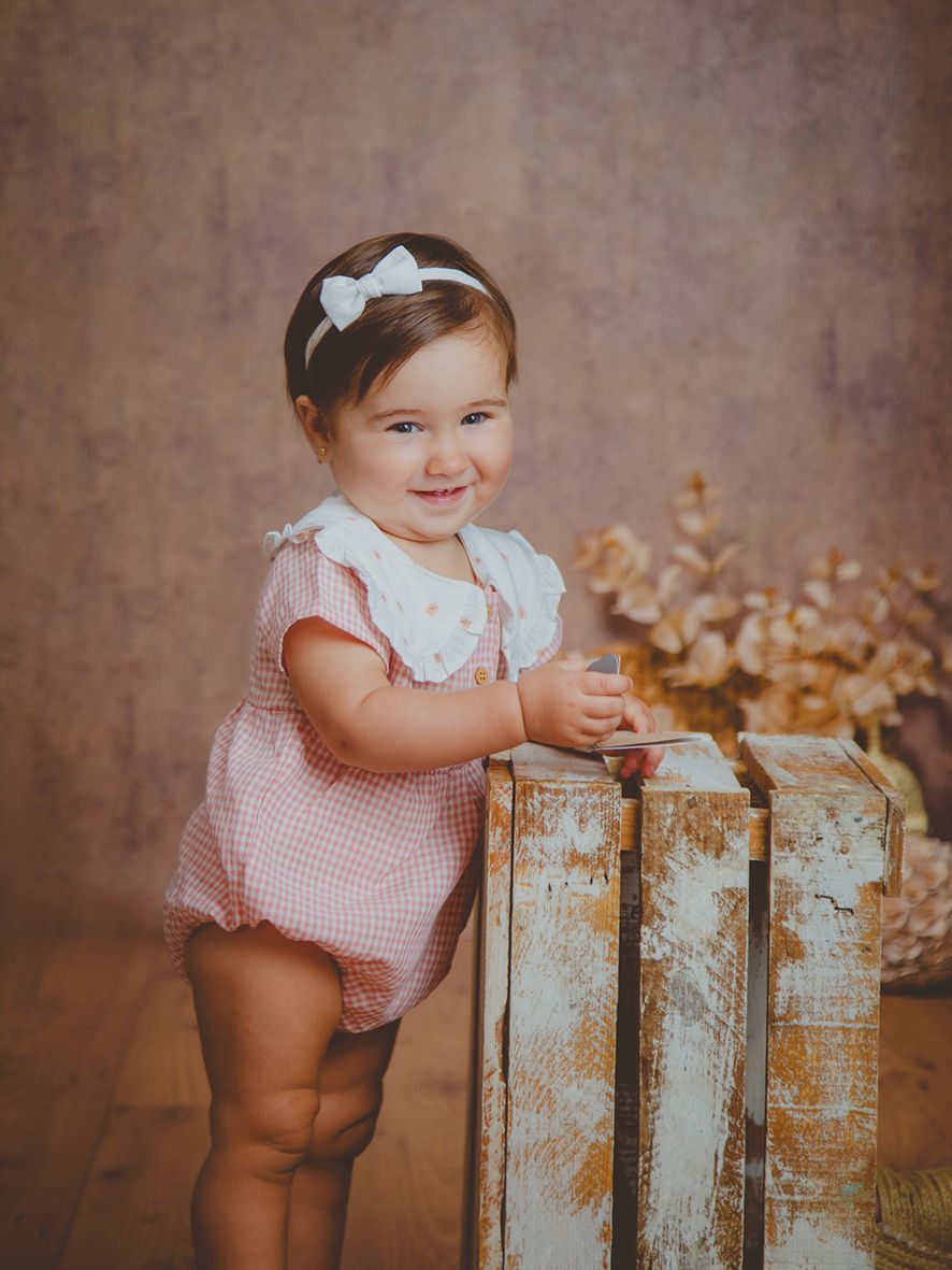 Dulce bebé con un pelele rosa y cuello blanco, de pie junto a una rústica caja de madera, sonriendo en su sesión de fotos en estudio.