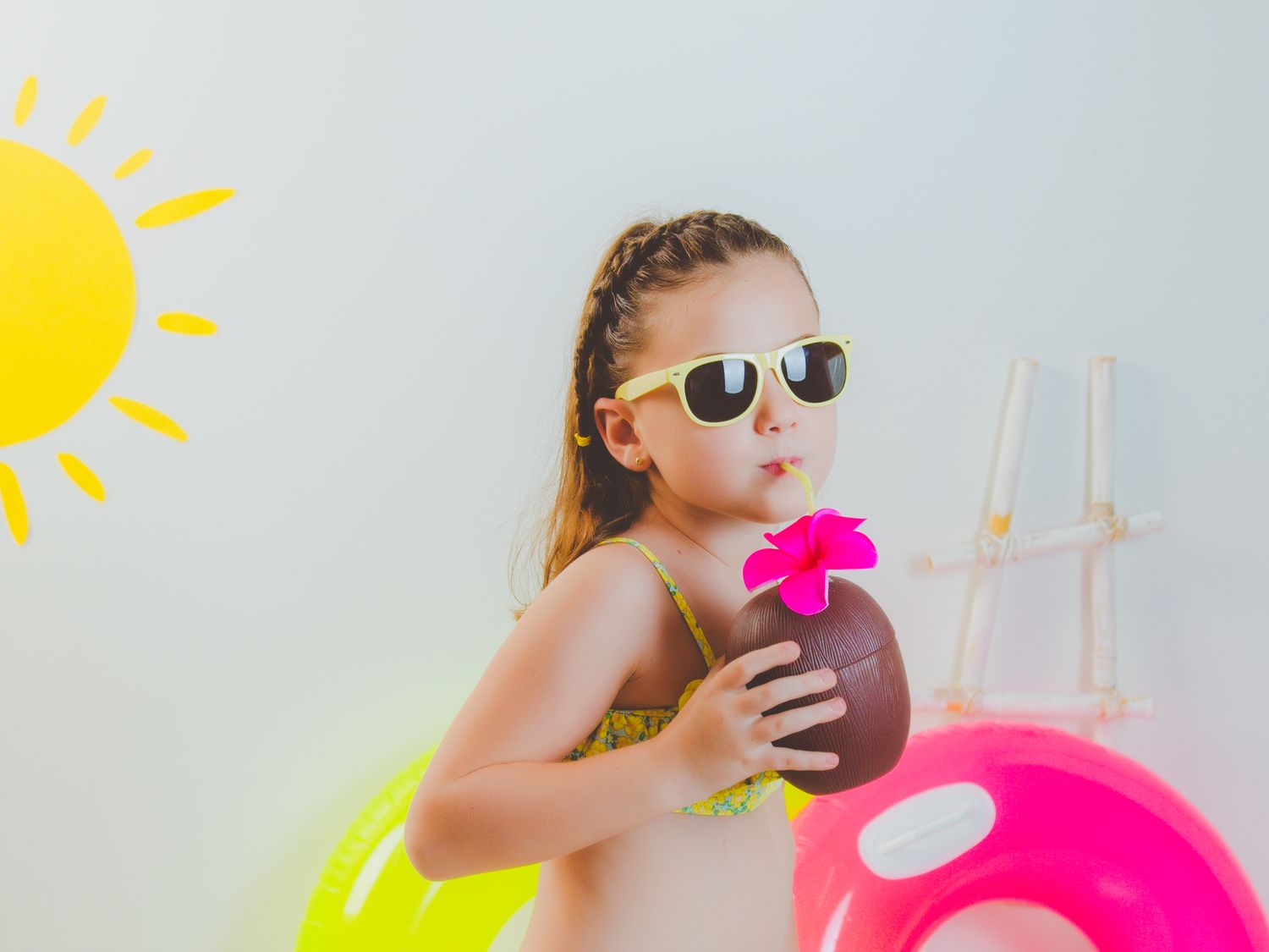 Niña en su sesión de verano, en traje de baño bebiendo de un refresco de coco, con gafas de sol amarillas y flotadores rosas y amarillos.