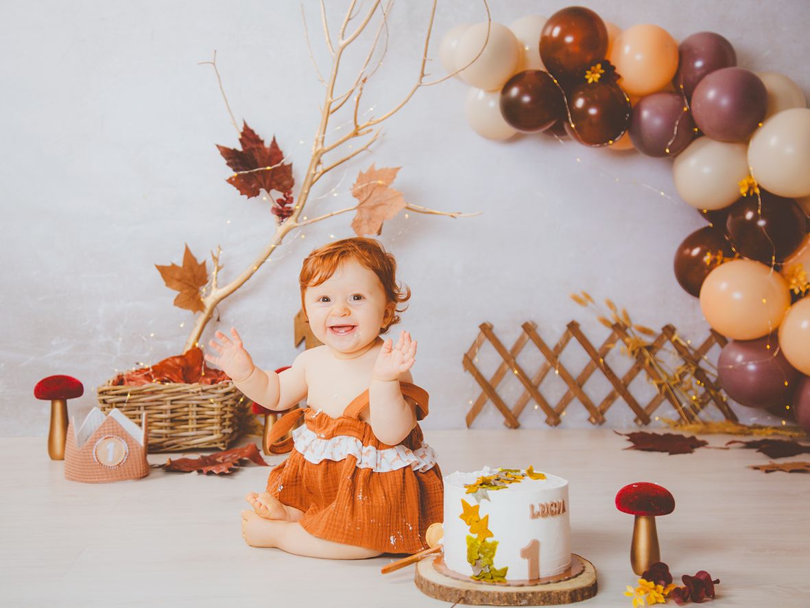 Una alegre bebé pelirroja con un vestido color óxido celebra su primer cumpleaños con una sesión de fotos con pastel con temática otoñal.