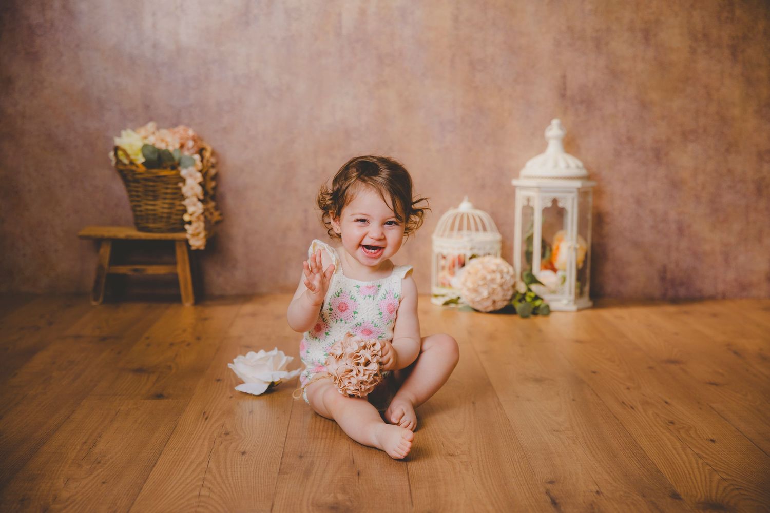Una niña pequeña, vestida con un pelele rosa, ríe alegremente sentada en un suelo de madera cerca de faroles decorativos.