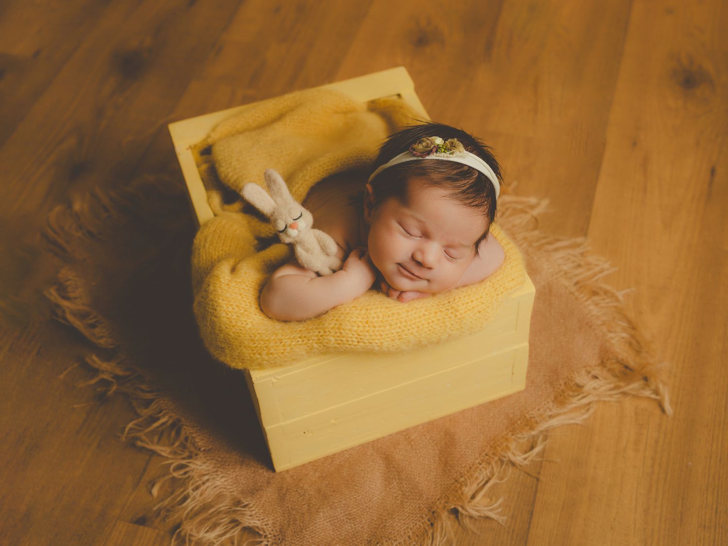 Fotografía de estudio de un recién nacido durmiendo en una caja de madera amarilla con un conejito abrazado sobre una suave manta amarilla.