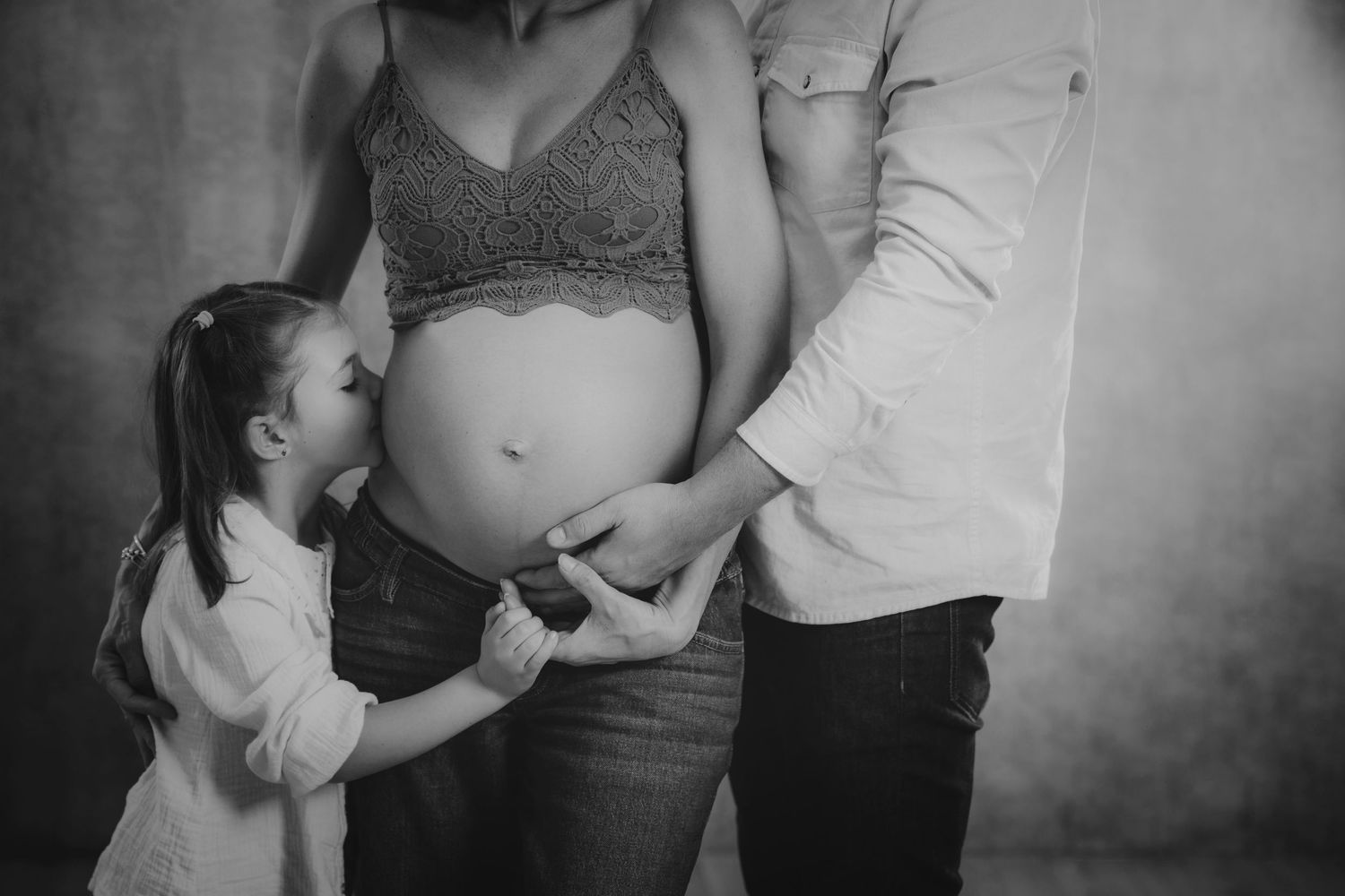 Fotografía en blanco y negro de una niña besando la barriga de su madre embarazada en un estudio.