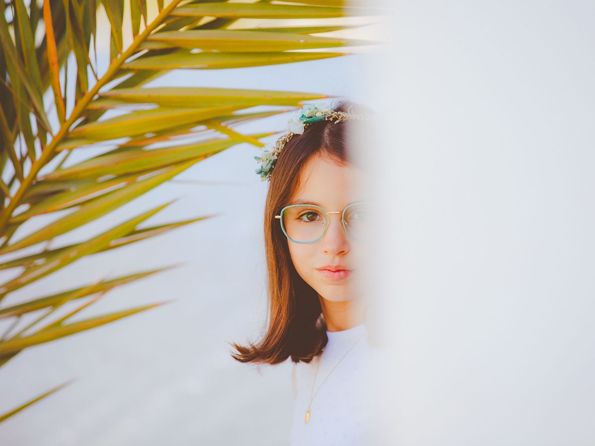 Retrato de comunión de exteriores en Almería de una niña con corona de flores y gafas se asoma por detrás de una pared blanca.