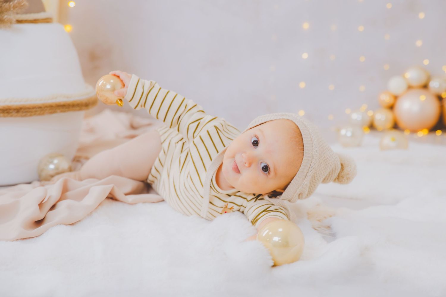 Bebé con pelele de rayas y gorro blanco, tumbado sobre una manta suave con adornos navideños dorados en su sesión de fotos de Navidad.