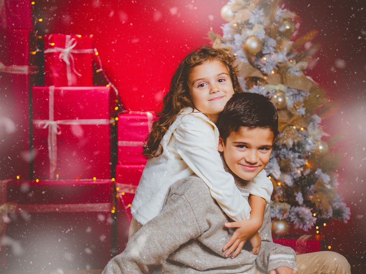 Hermanos posan abrazados en un decorado de Navidad con fondo rojo de cajas de regalo y árbol navideño en tonos dorados y blanco.