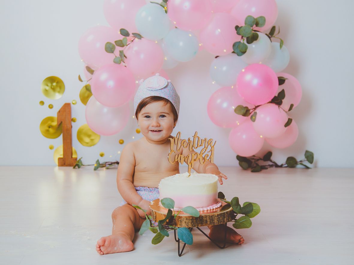 Niña en su sesión de fotos de primer cumpleaños con pastel blanco y corona de tela rodeada de decoración de globos rosas y blancos.