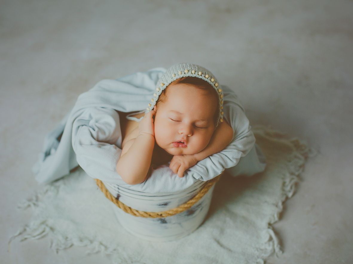 Bebé recién nacido durmiendo, con un gorro, acurrucado en un cubo de madera blanco con una tela celeste suave.