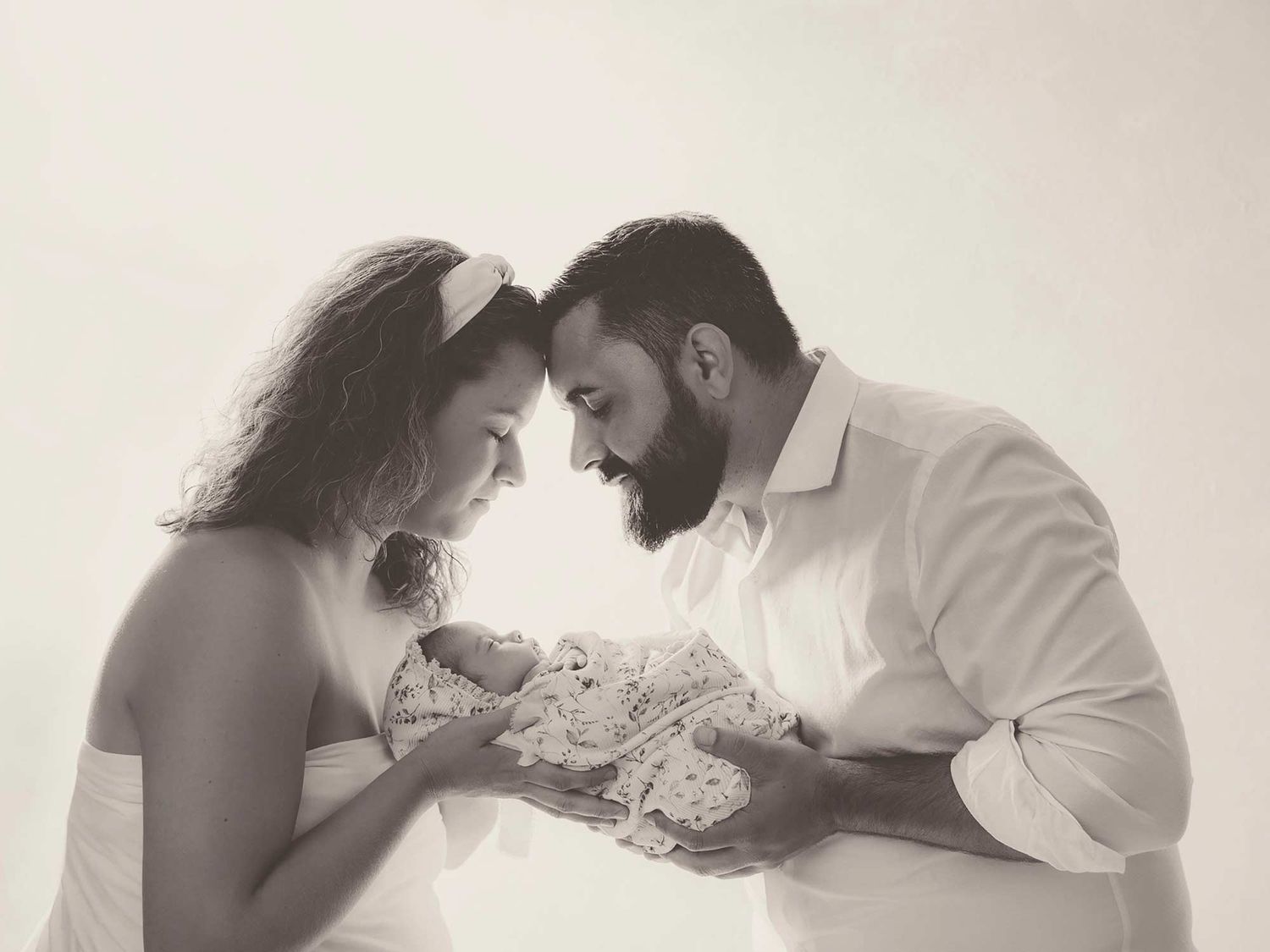 Black and white photo of parents tenderly gazing at their newborn baby in a bright studio setting.
