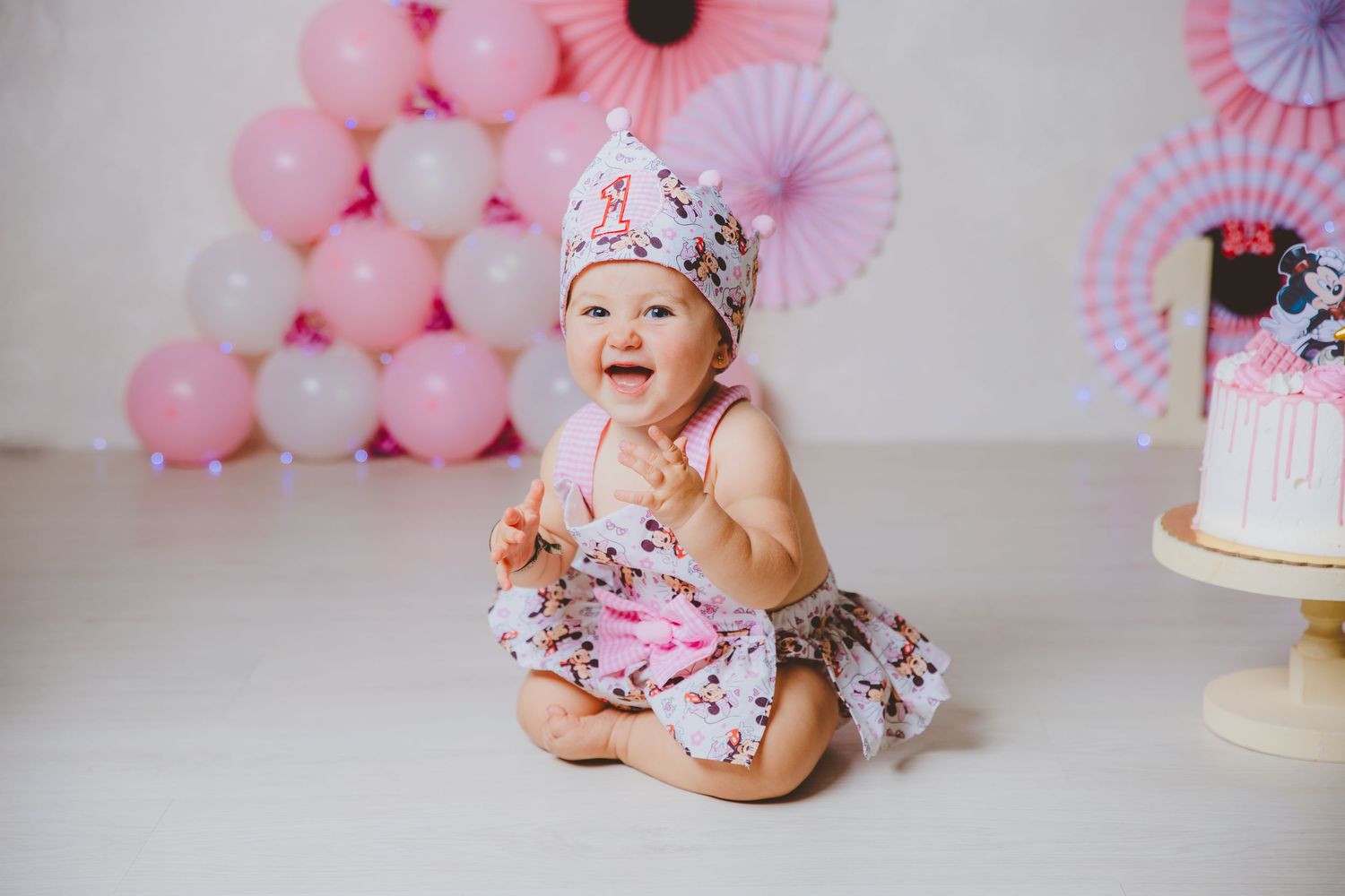 Una niña pequeña, vestida con un traje rosa de ratoncita con lazo, se ríe durante la sesión de fotos de su primer cumpleaños con pastel.