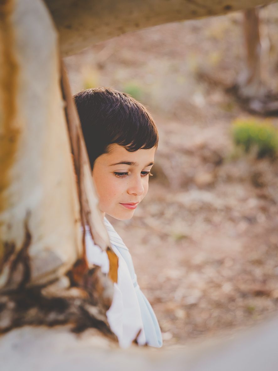 Fotografía de perfil de un niño de comunión  apoyado en un tronco de un árbol.