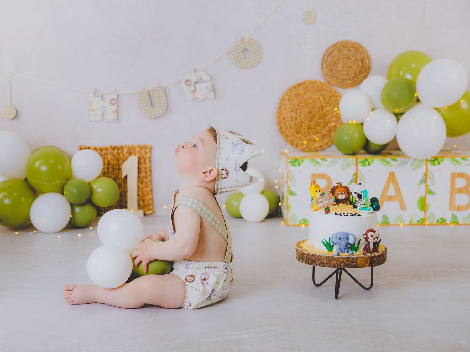 Niño sentado junto a su tarta en su sesión de primer cumpleaños rodeado de globos y decoración con temática de animalitos.