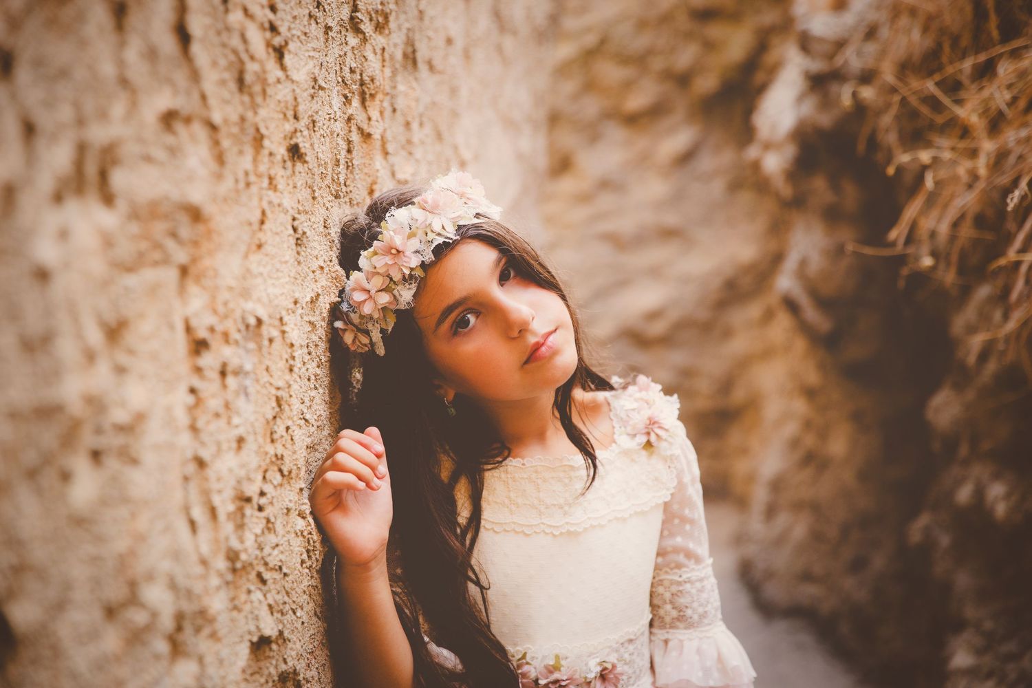 Preciosa niña con vestido de comunión y tocado con flores apoyada contra un muro de piedra.