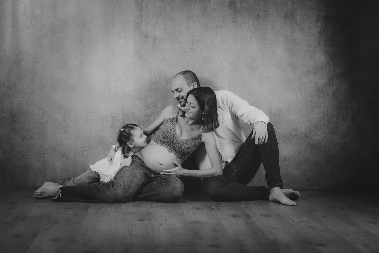 Fotografía de maternidad en blanco y negro de una pareja y su hija pequeña sentados en el suelo de un estudio.