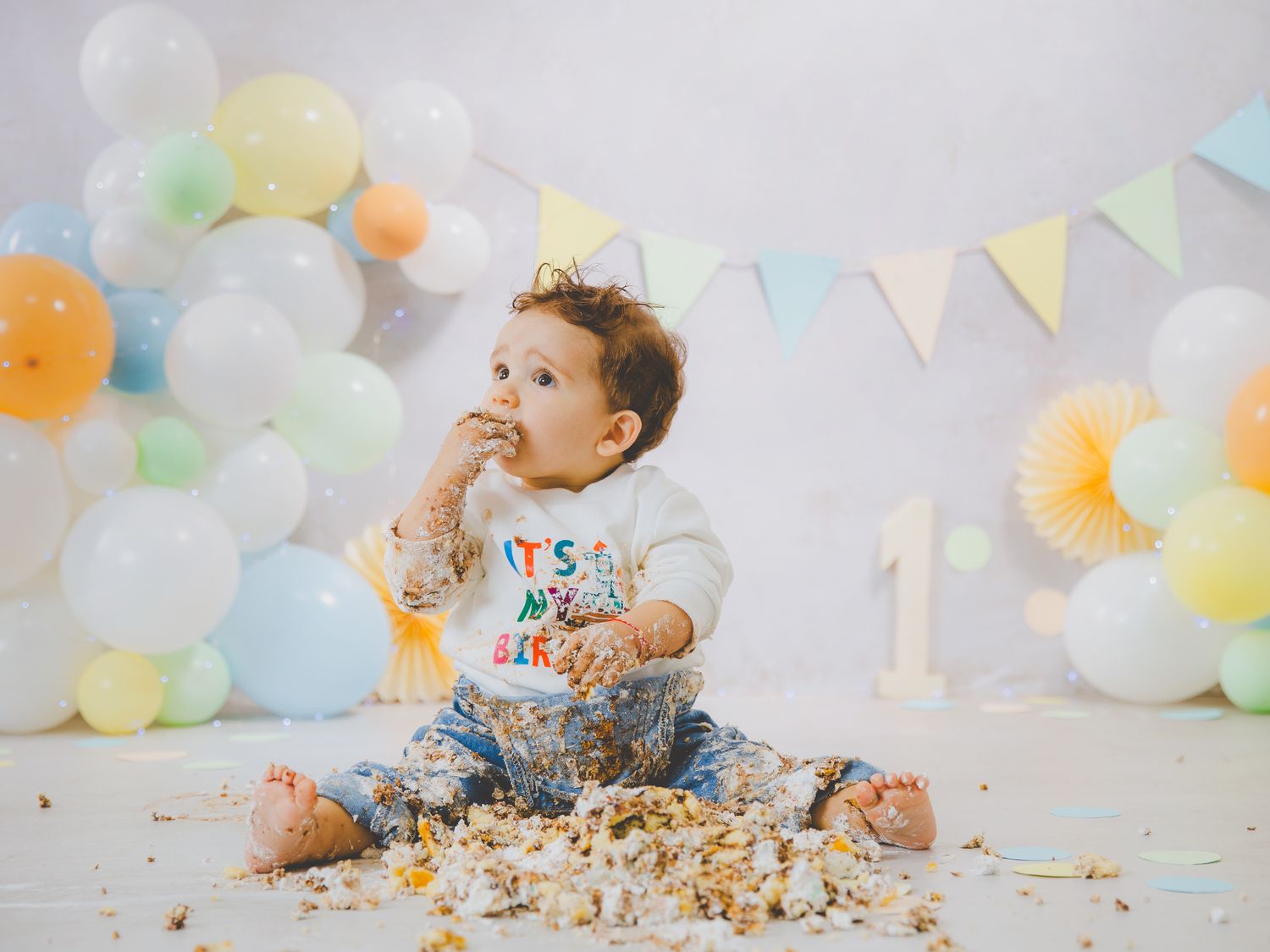 Bebé en su sesión de primer cumpleaños comiendo pastel destrozado, rodeado de decoración de globos en tonos pastel.