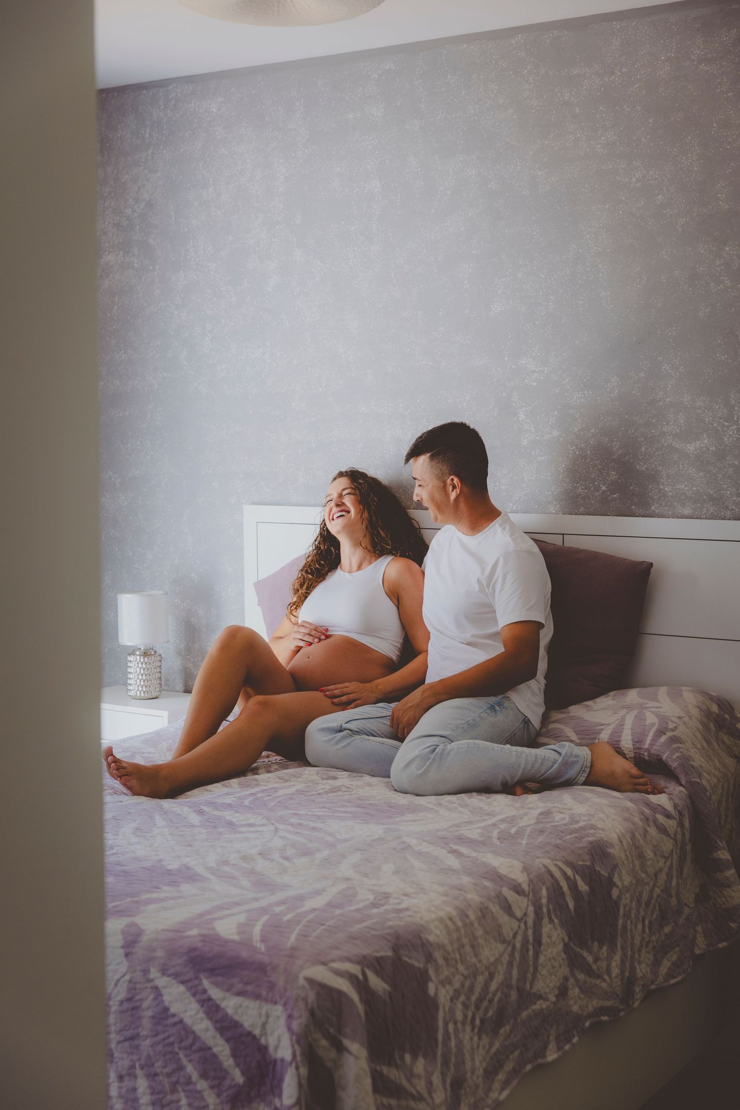 Una mujer embarazada sonriente y su pareja están sentados juntos en una cama, acariciando con cariño su vientre, durante su reportaje.
