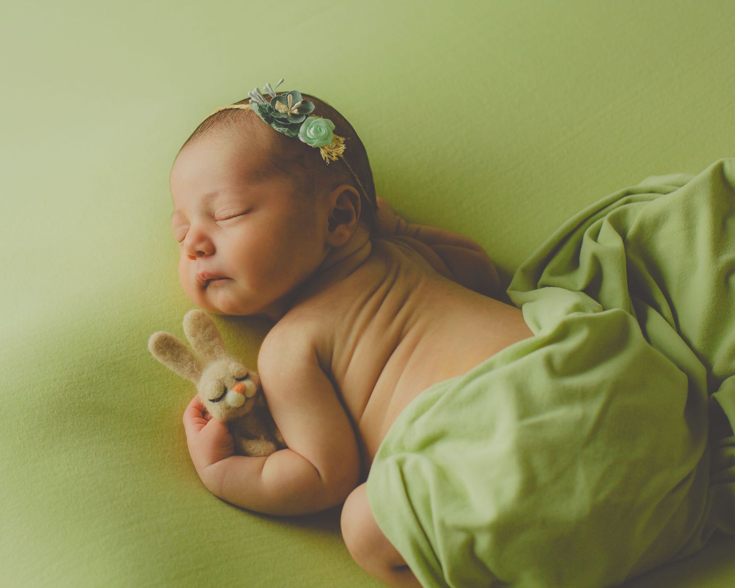 Recién nacido durmiendo durante su reportaje de fotos, con una diadema de flores, abrazando un conejito de peluche en una tela suave verde.
