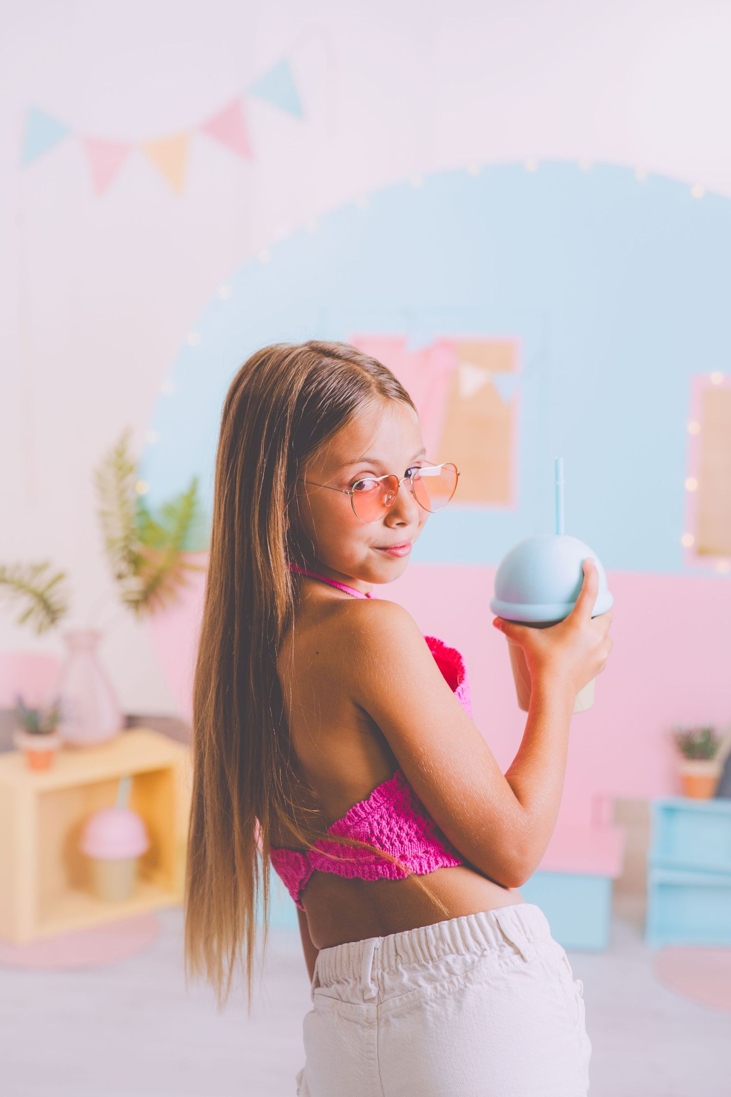 Una niña rubia con un top corto rosa sostiene un vaso en forma de helado mientras posa en su sesión de verano.