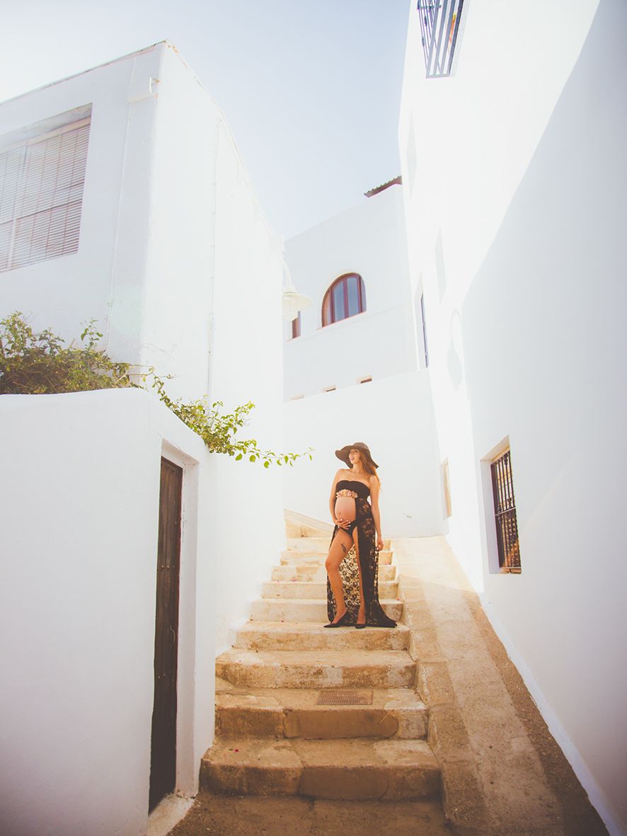 Embarazada posando en una escalera con sombrero y vestido negro en un pueblo mediterráneo como es Agua Amarga.