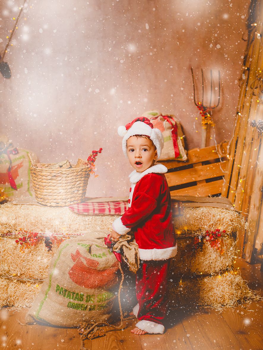 Un niño pequeño con gesto de sorprendido y vestido de Papá Noel posa junto a un saco de arpillera en un decorado navideño rústico y nevado.