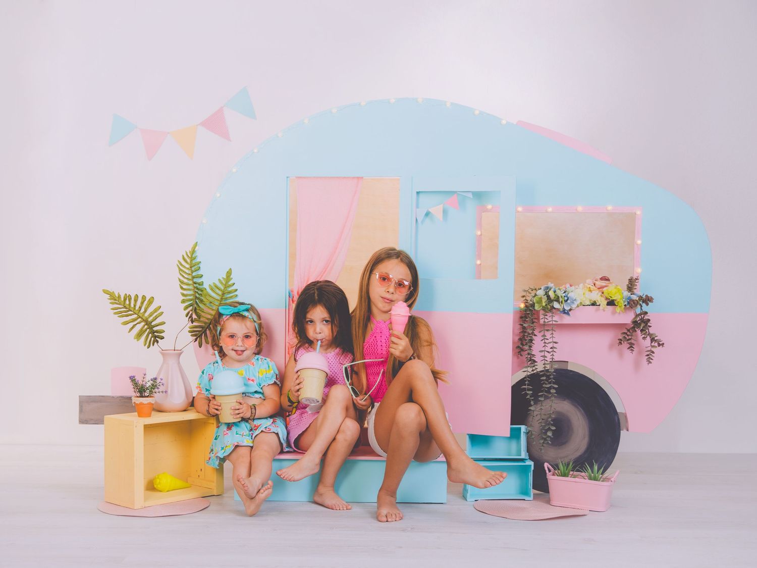Tres niñas sentadas posando frente al decorado de verano con una caravana en tonos pastel, en su sesión de fotografías.