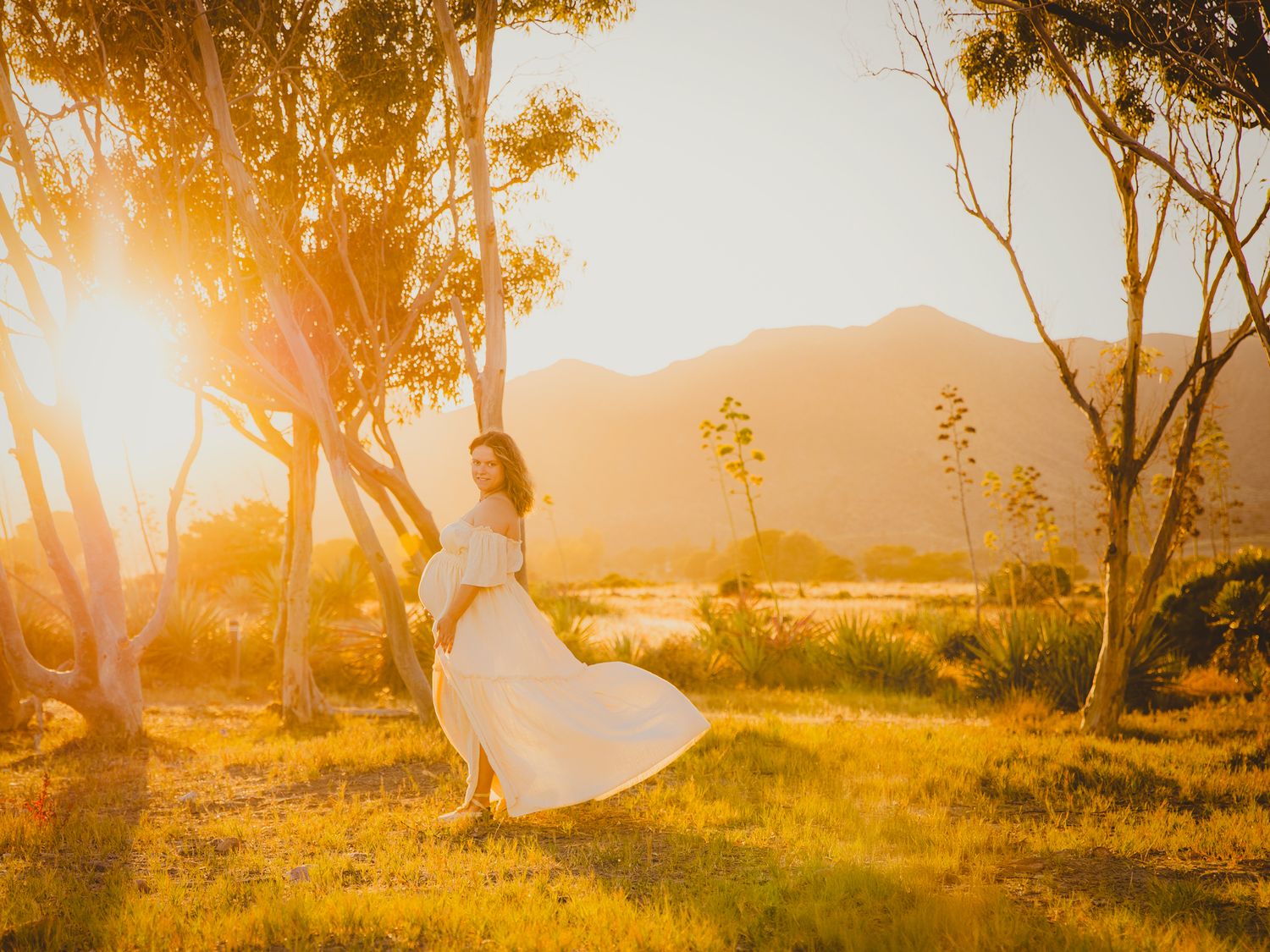 Embarazada con un vestido vaporoso camina en una pradera dorada bañada por el sol y arbolado en su sesión de exteriores en Cabo de Gata.